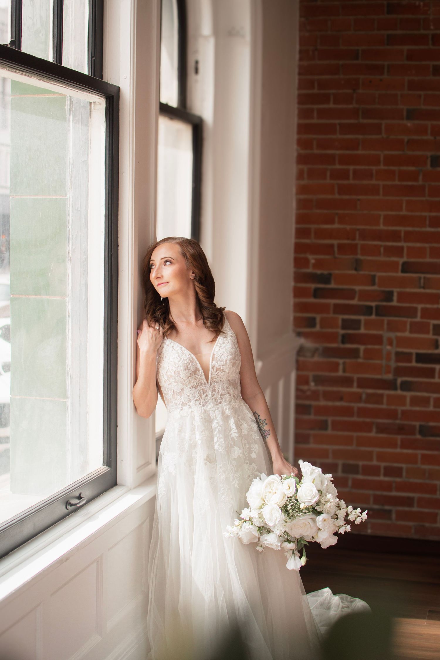 Bride in lace wedding gown holding white bouquet gazes out large window beside exposed brick wall.