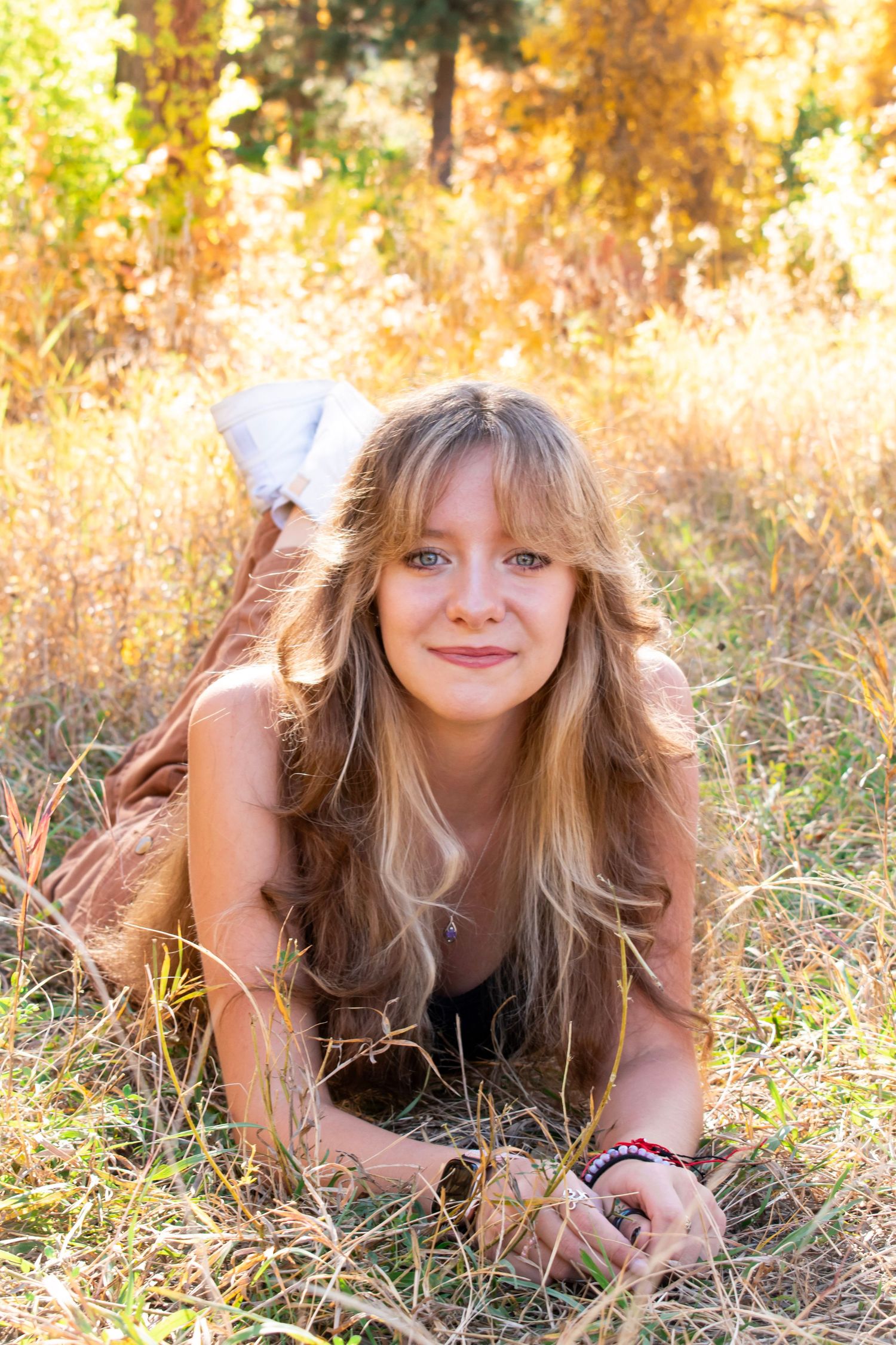 Young blonde woman with long hair lying in golden dry grass field, smiling at camera in warm sunlight.