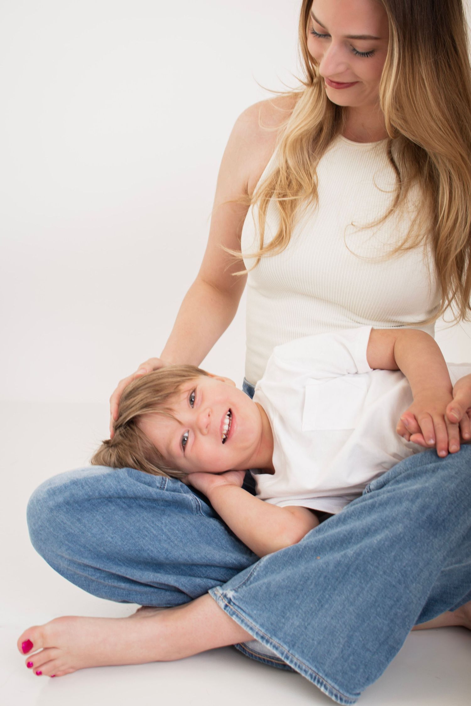 A smiling toddler rests in a young woman's crossed legs on a white background, sharing a joyful moment together.