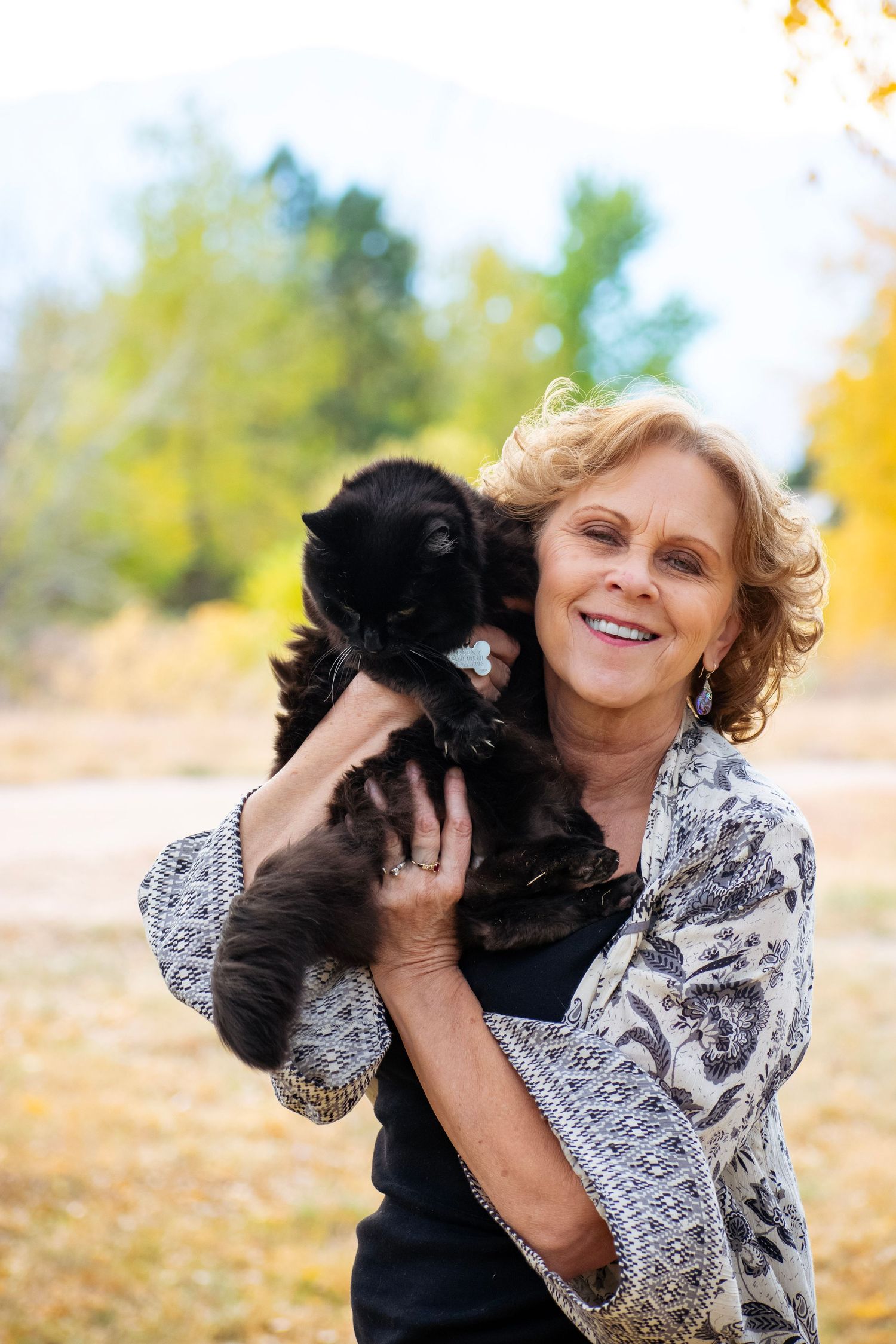 Smiling woman with blonde hair holds a black cat outdoors among autumn yellow trees.