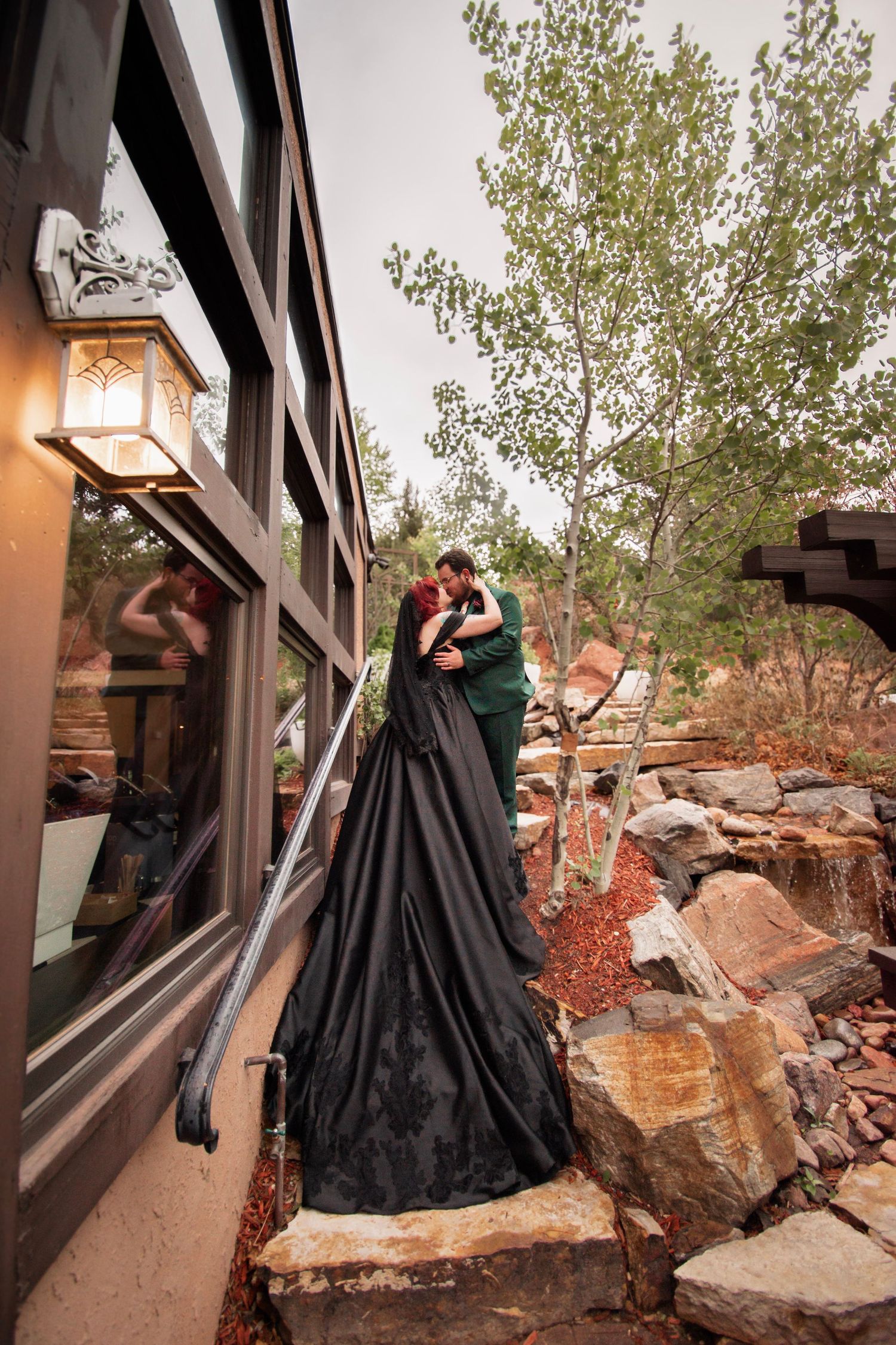 Couple embracing in black and green wedding attire on rocky steps beside a rustic building surrounded by trees.