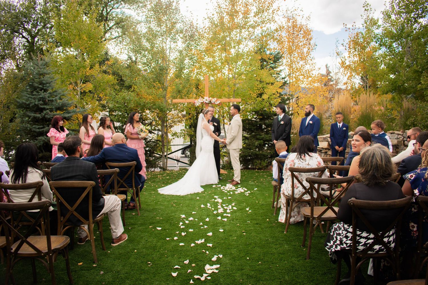 Outdoor fall wedding ceremony on green lawn with couple at altar, guests seated in wooden chairs, colorful autumn trees in background.