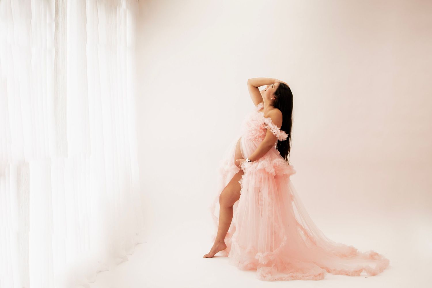 Pregnant woman in a flowing pink tulle maternity gown poses elegantly in a bright, soft-lit studio beside sheer white curtains.
