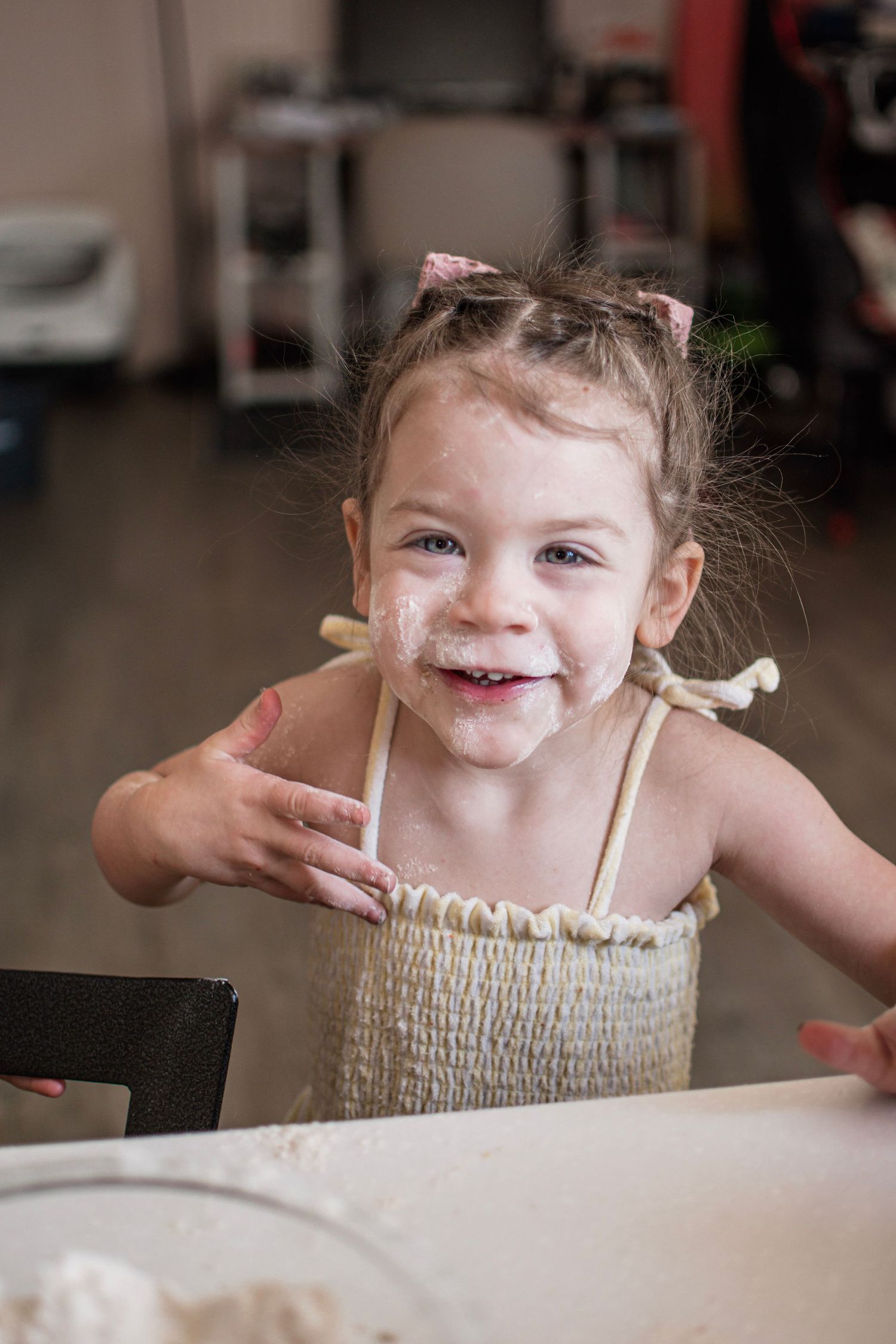 Happy toddler girl with flour on her face smiling while helping bake in the kitchen wearing a woven apron.