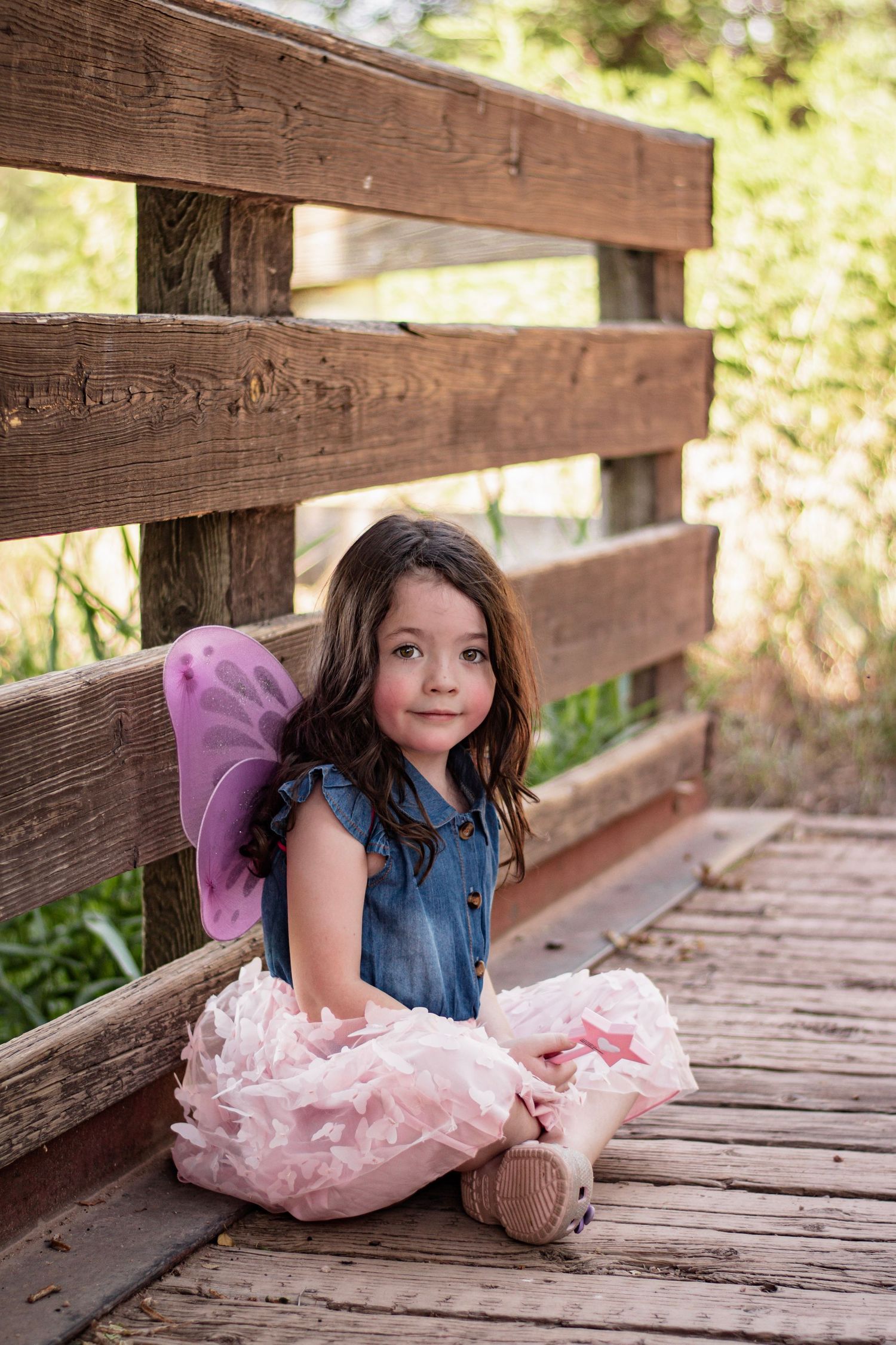 A young girl in a denim dress, pink tutu, and fairy wings sits cross-legged on a wooden bridge outdoors.