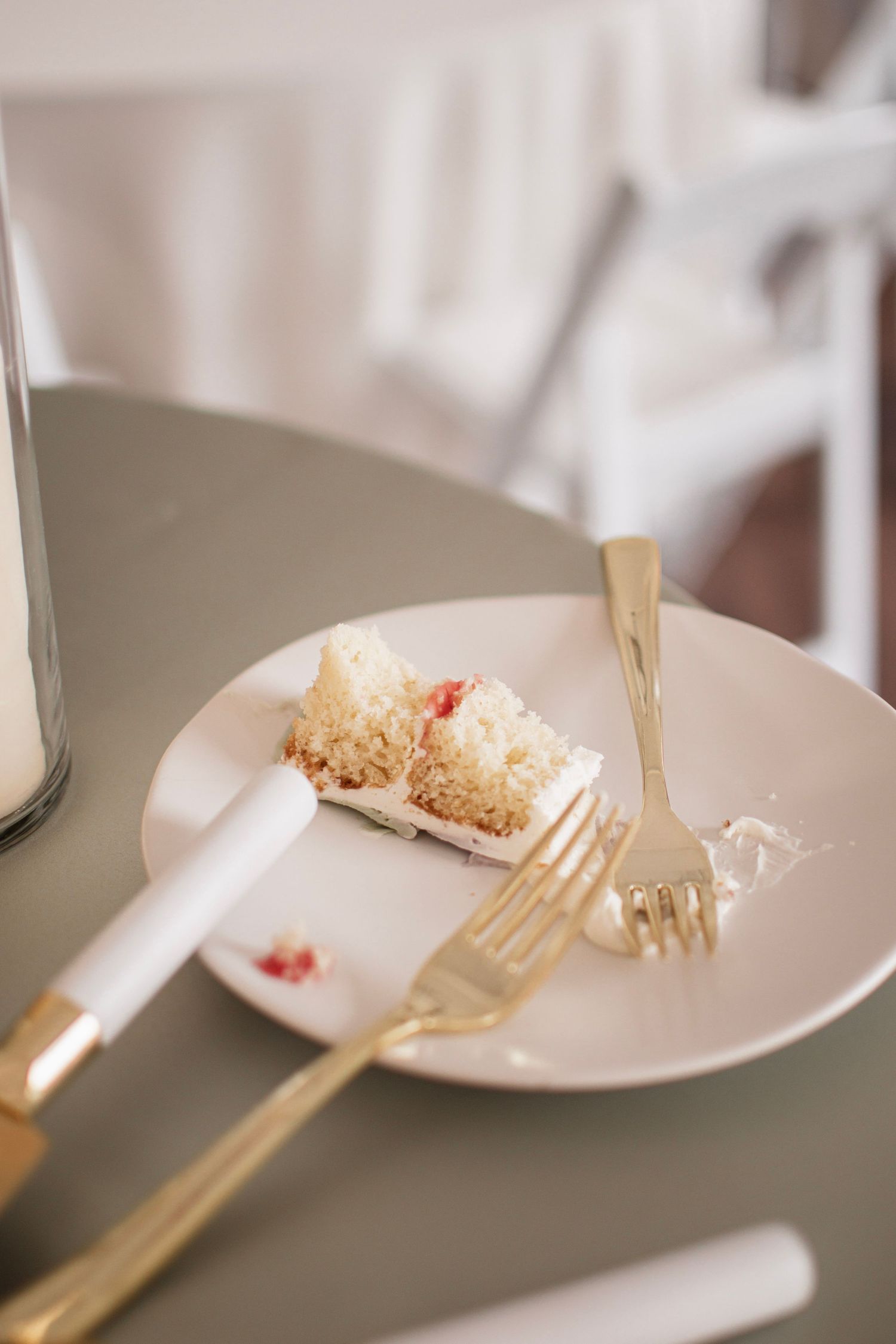 A partially eaten slice of white cake with strawberry filling on a pink plate with gold fork and knife on a gray table.
