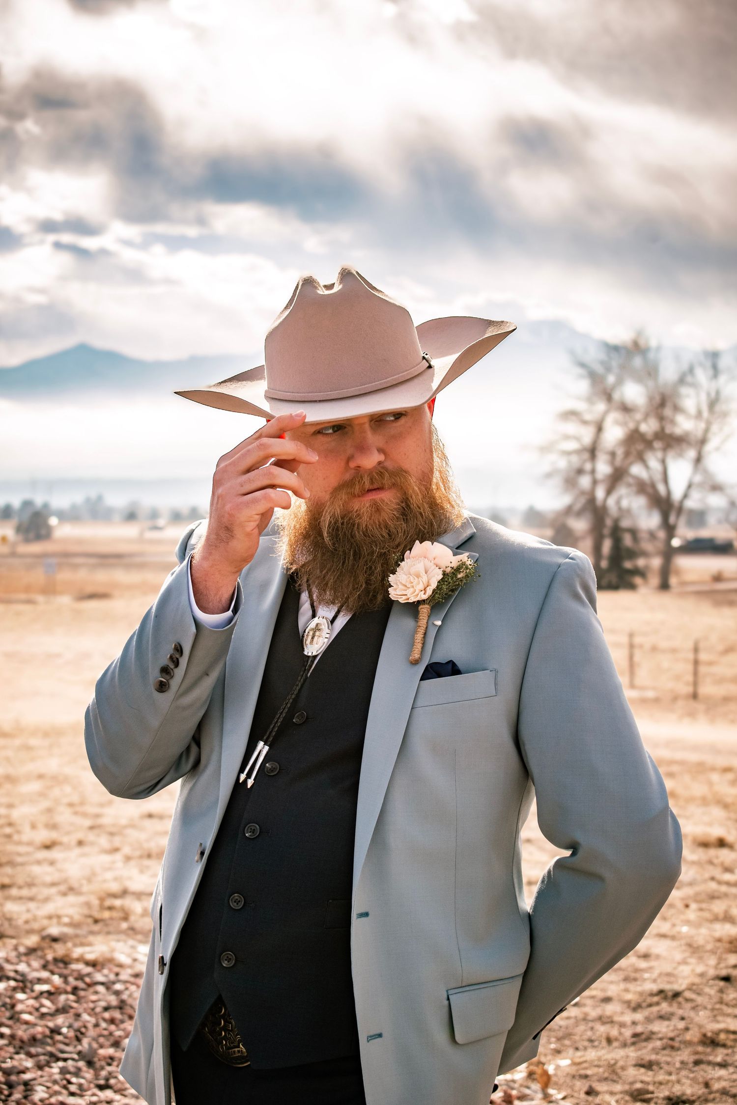 Bearded groom in light gray suit and cowboy hat tips brim outdoors at rustic ranch wedding venue.