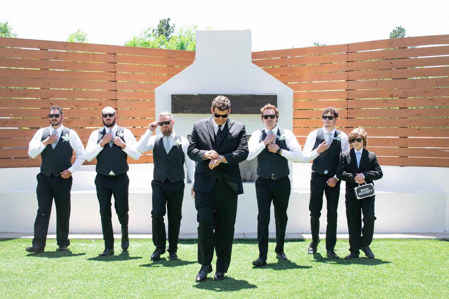Groom and groomsmen in black suits and sunglasses posing outdoors at a wedding venue on a sunny day.