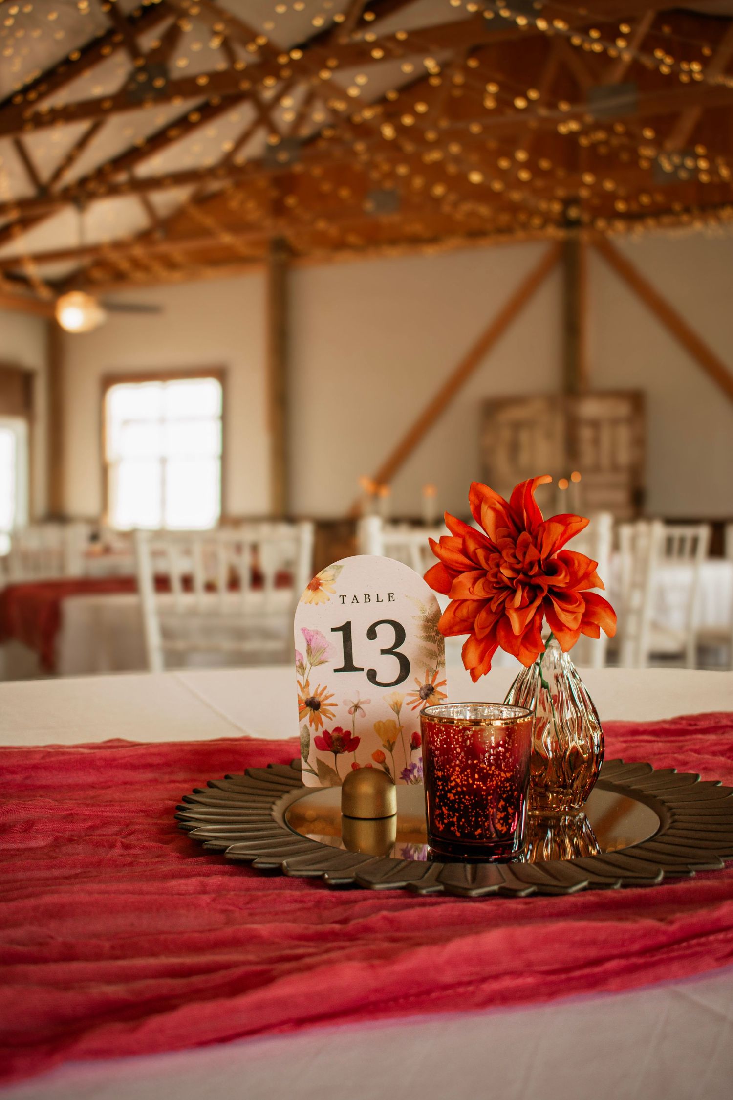 Elegant wedding reception table 13 centerpiece with red dahlia flower, jeweled glasses, and string lights in rustic barn venue.