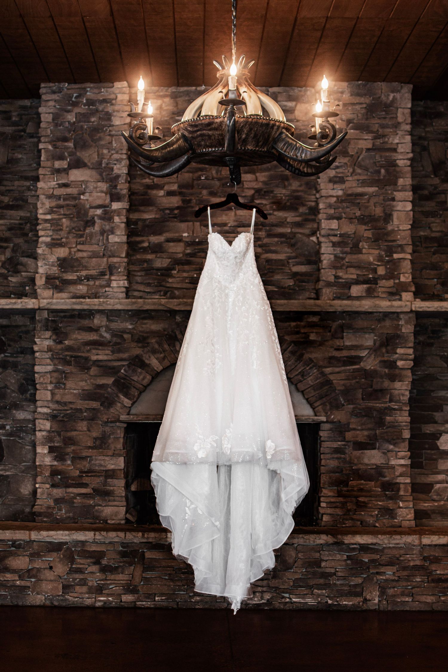Elegant white lace wedding dress hanging from a rustic chandelier in front of a stone fireplace.