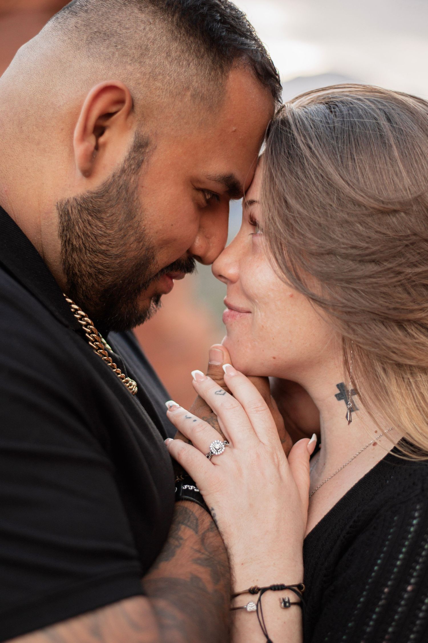 Romantic couple sharing an intimate moment, touching foreheads, woman displaying engagement ring, both wearing black outfits.