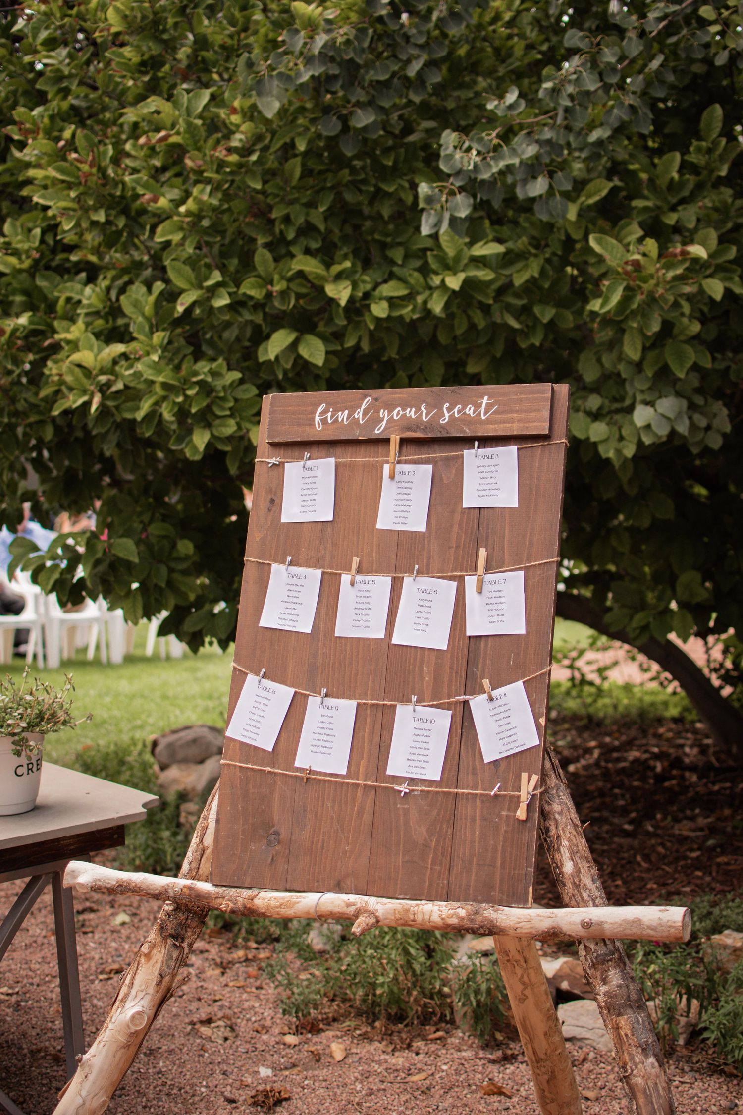 Rustic wooden wedding seating chart display with twine and clothespins, propped on log easel outdoors near green trees.