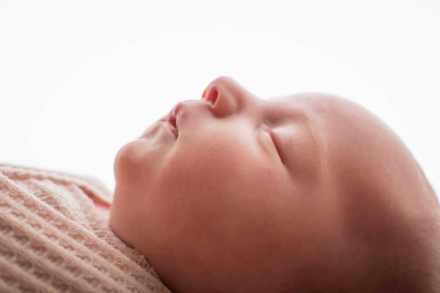 Peaceful newborn baby sleeping soundly, wrapped in a soft pink knit blanket, photographed against a white background.