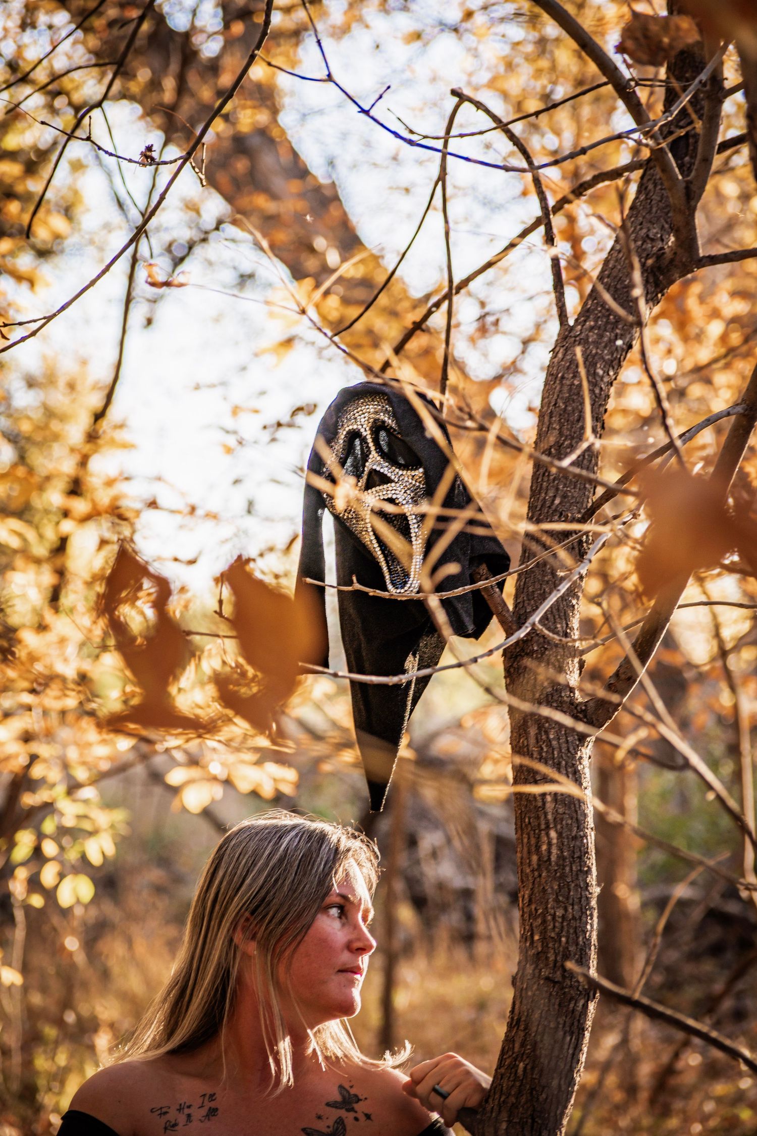 Woman with tattoos gazes up at a Scream mask hanging from autumn tree branches in a golden fall forest setting.