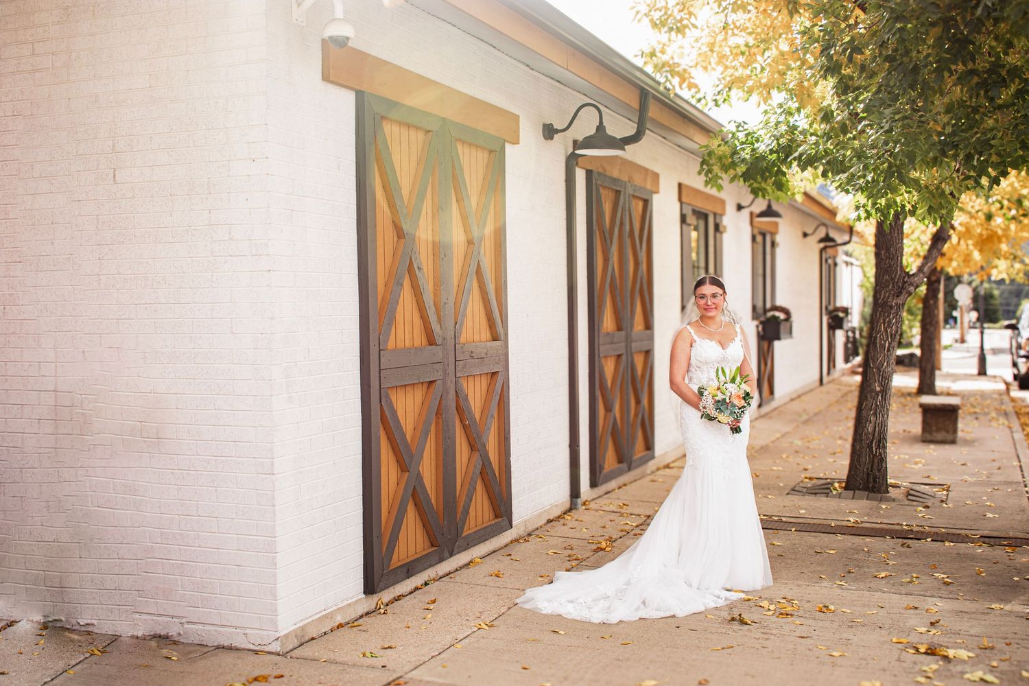 Bride in white wedding gown poses outside rustic barn with wooden doors and brick walls on a sunny autumn day.