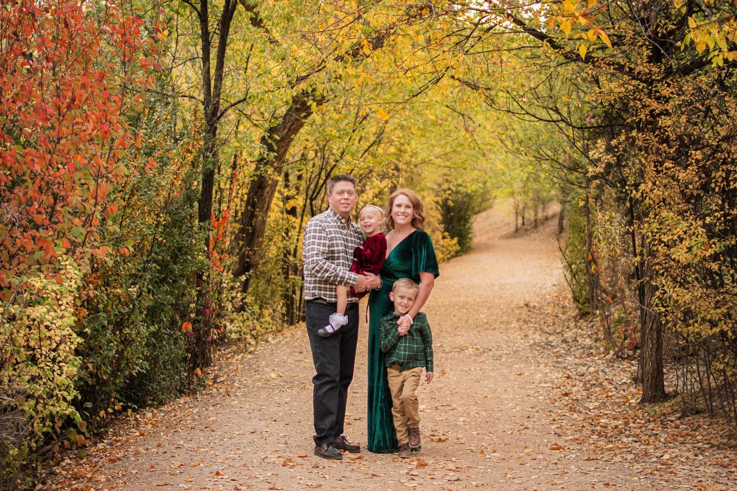 Family of four poses together on a tree-lined dirt path surrounded by colorful orange and yellow autumn foliage.