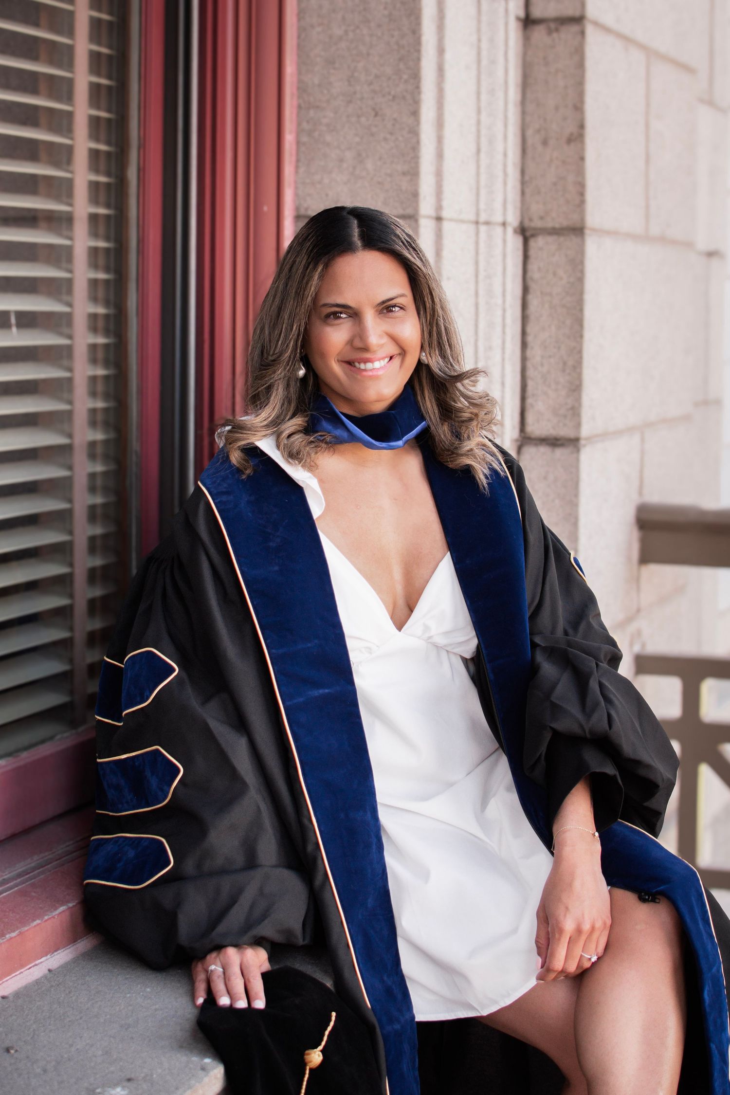 Smiling woman in black and blue doctoral graduation regalia sitting outdoors on steps near a brick building.
