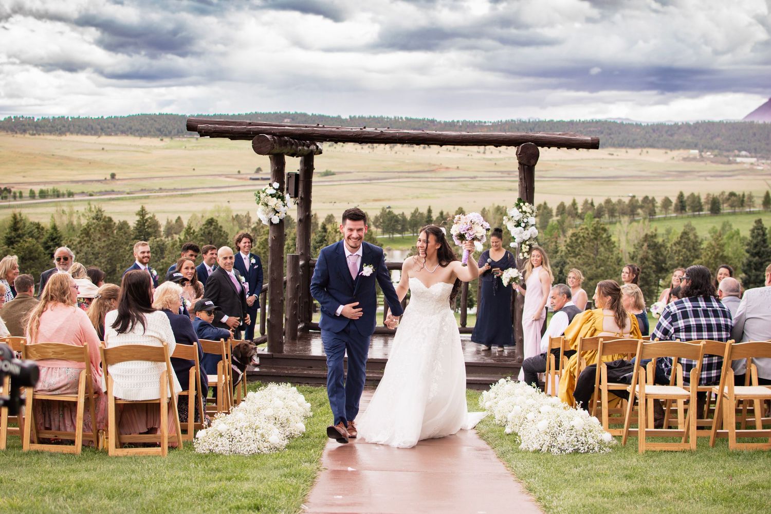 Bride and groom walk down outdoor aisle after wedding ceremony with scenic mountain meadow backdrop.