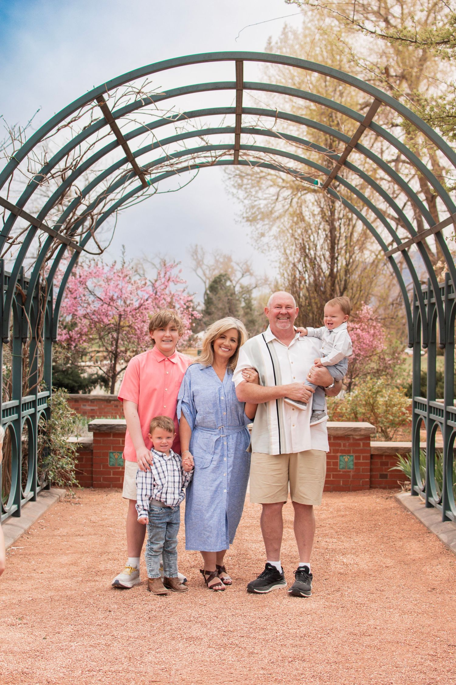 Family of five posing under a green metal garden arch tunnel with spring blossoms in the background.