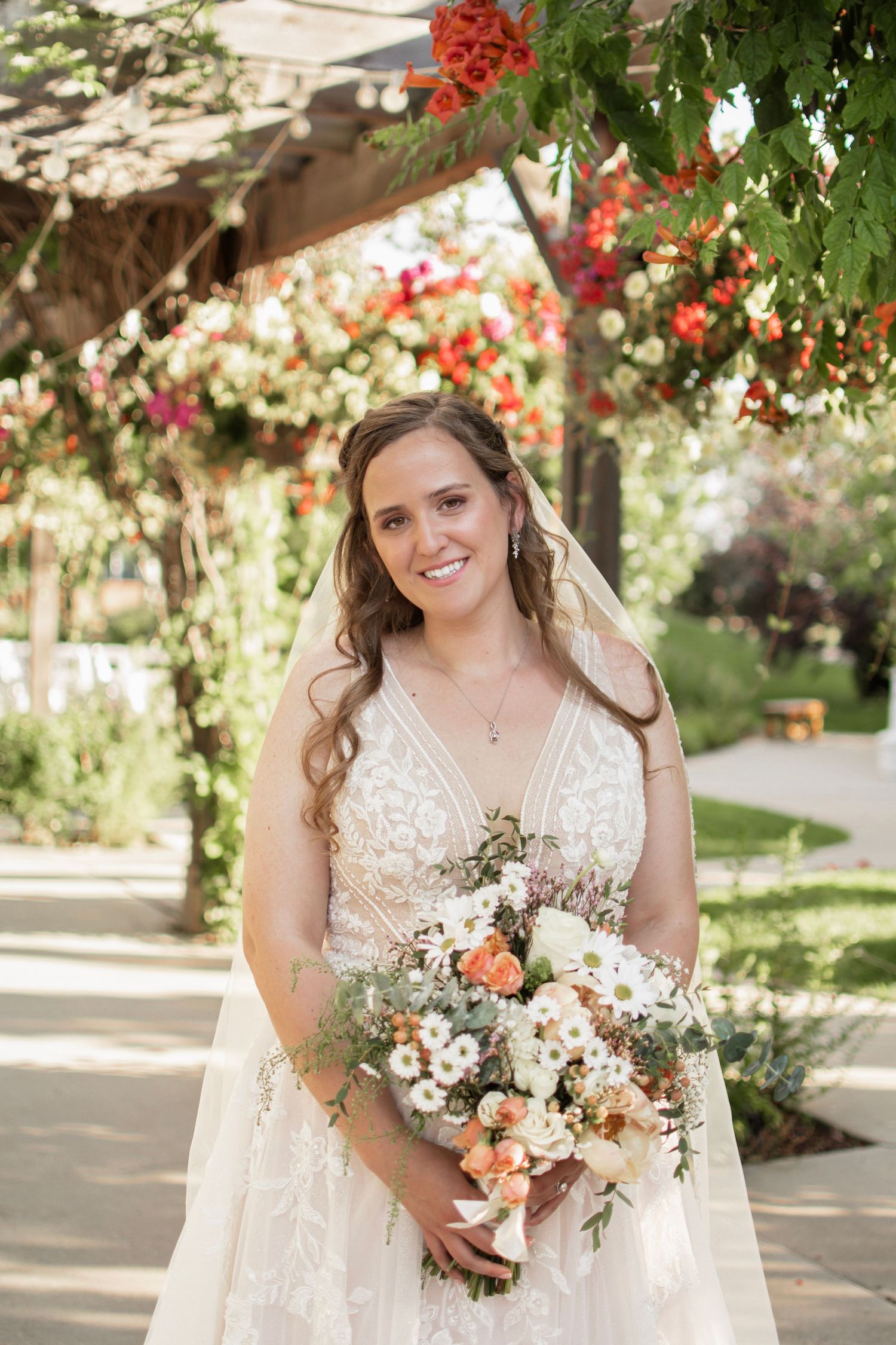 Smiling bride in lace gown holding peach and white bouquet under floral pergola outdoors.