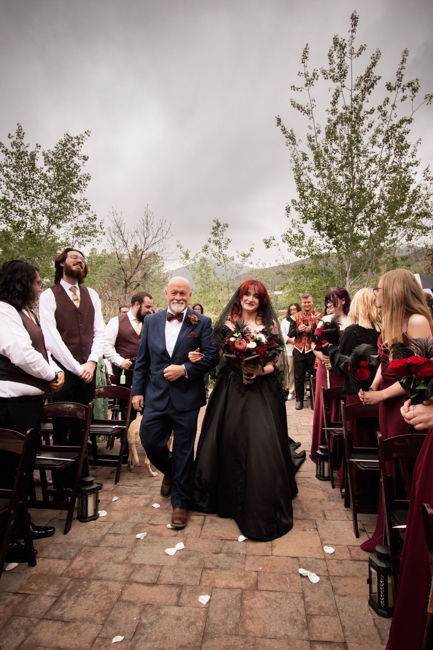 Bride in black gown walks down outdoor aisle with escort, flanked by guests in burgundy and navy attire.