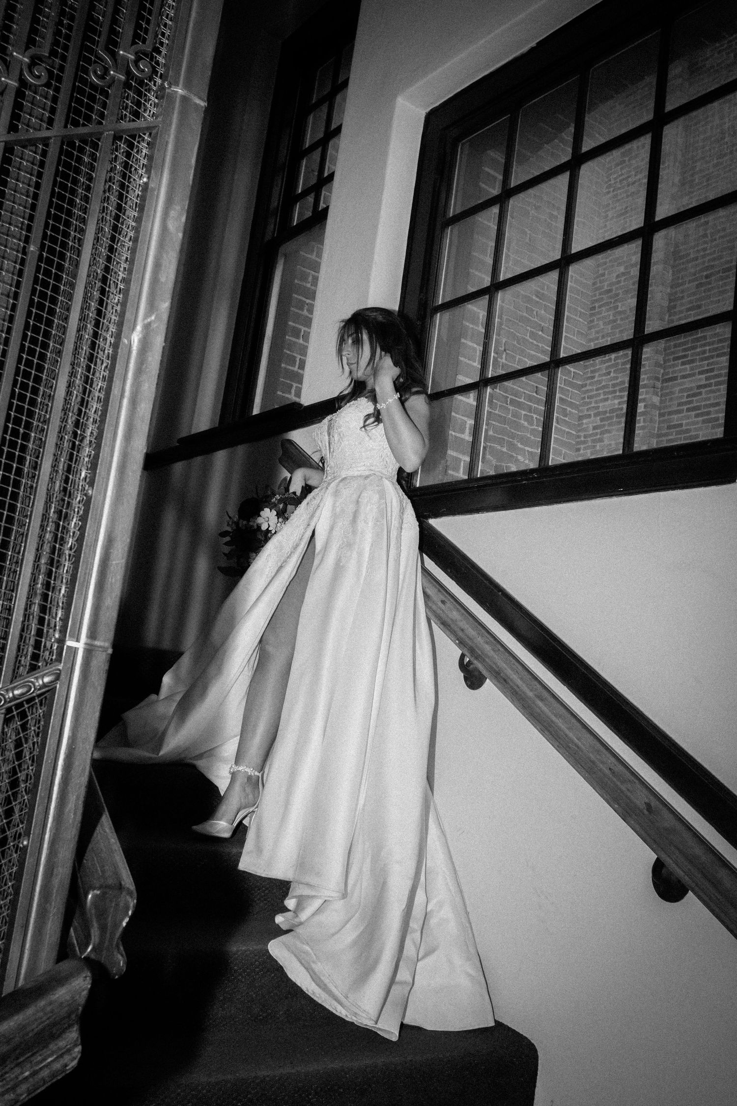 Black and white photo of a bride in a flowing white gown ascending a staircase near a large window.