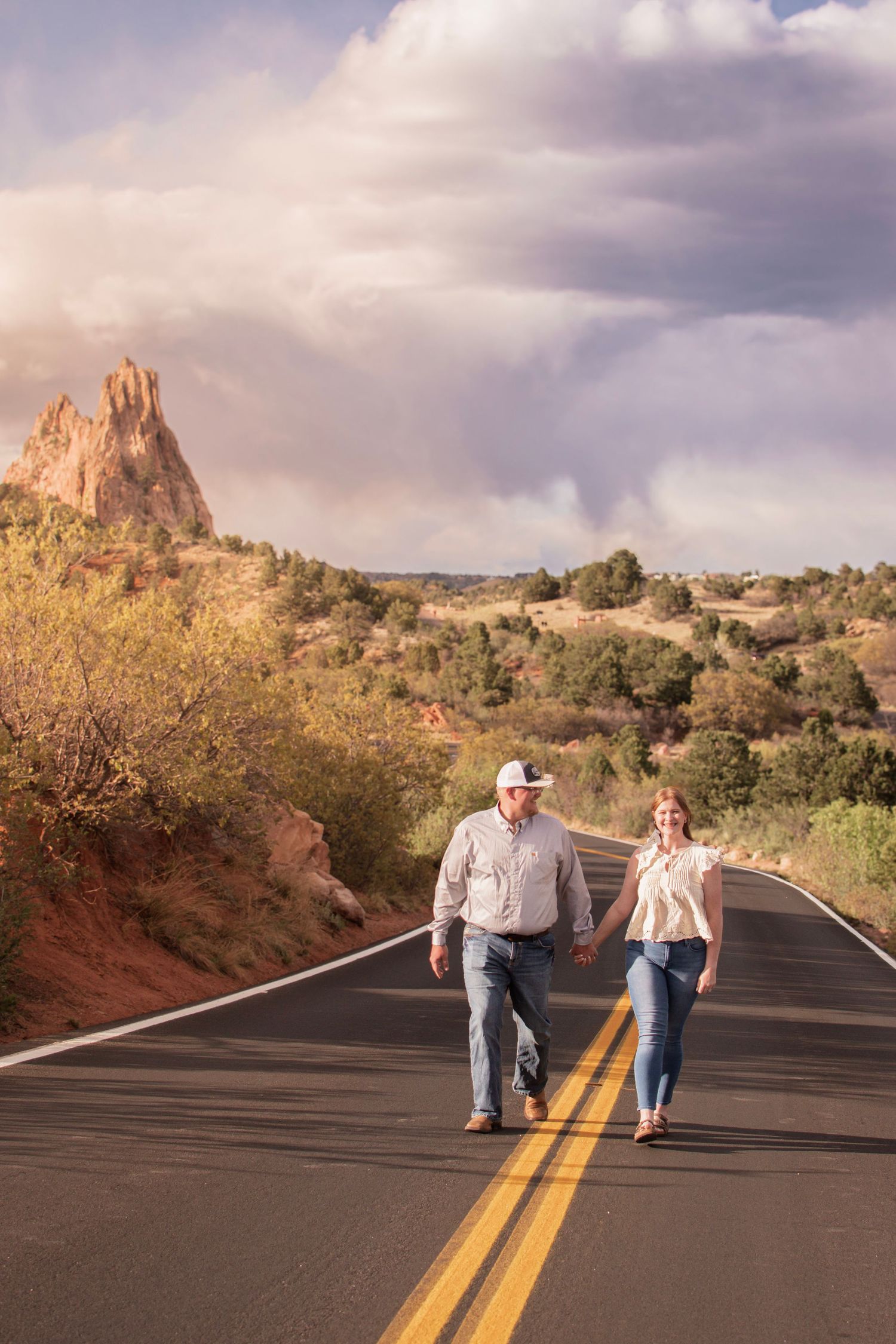 Couple holding hands walking on a scenic road at Garden of the Gods, Colorado, with red rock formations and dramatic sky.