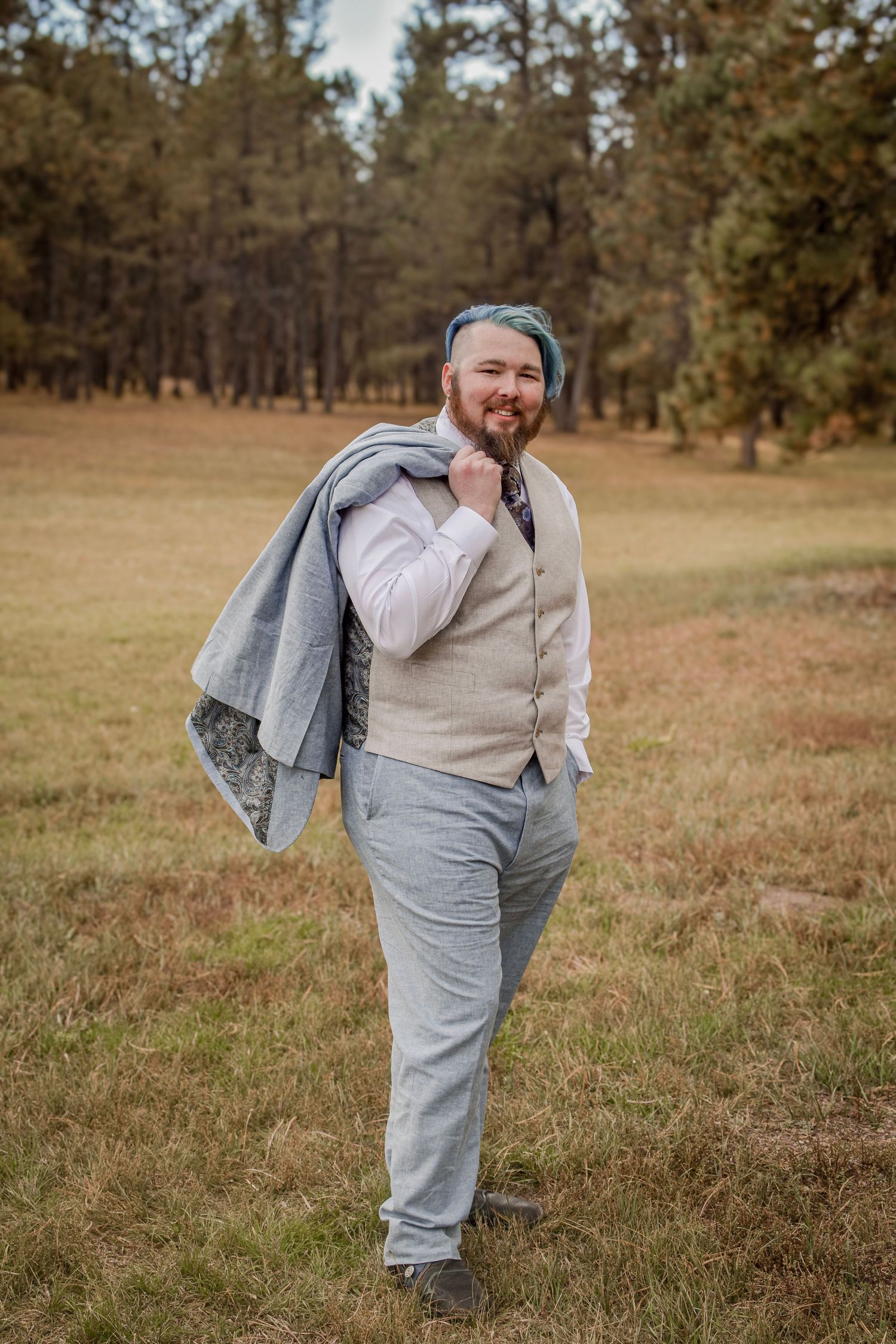 Smiling bearded man with blue hair in gray vest and trousers carrying jacket over shoulder in outdoor field.