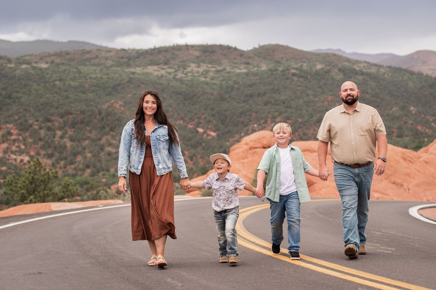 Family of four holding hands walking on a scenic mountain road with red rock formations and green hills backdrop at Garden of the Gods.