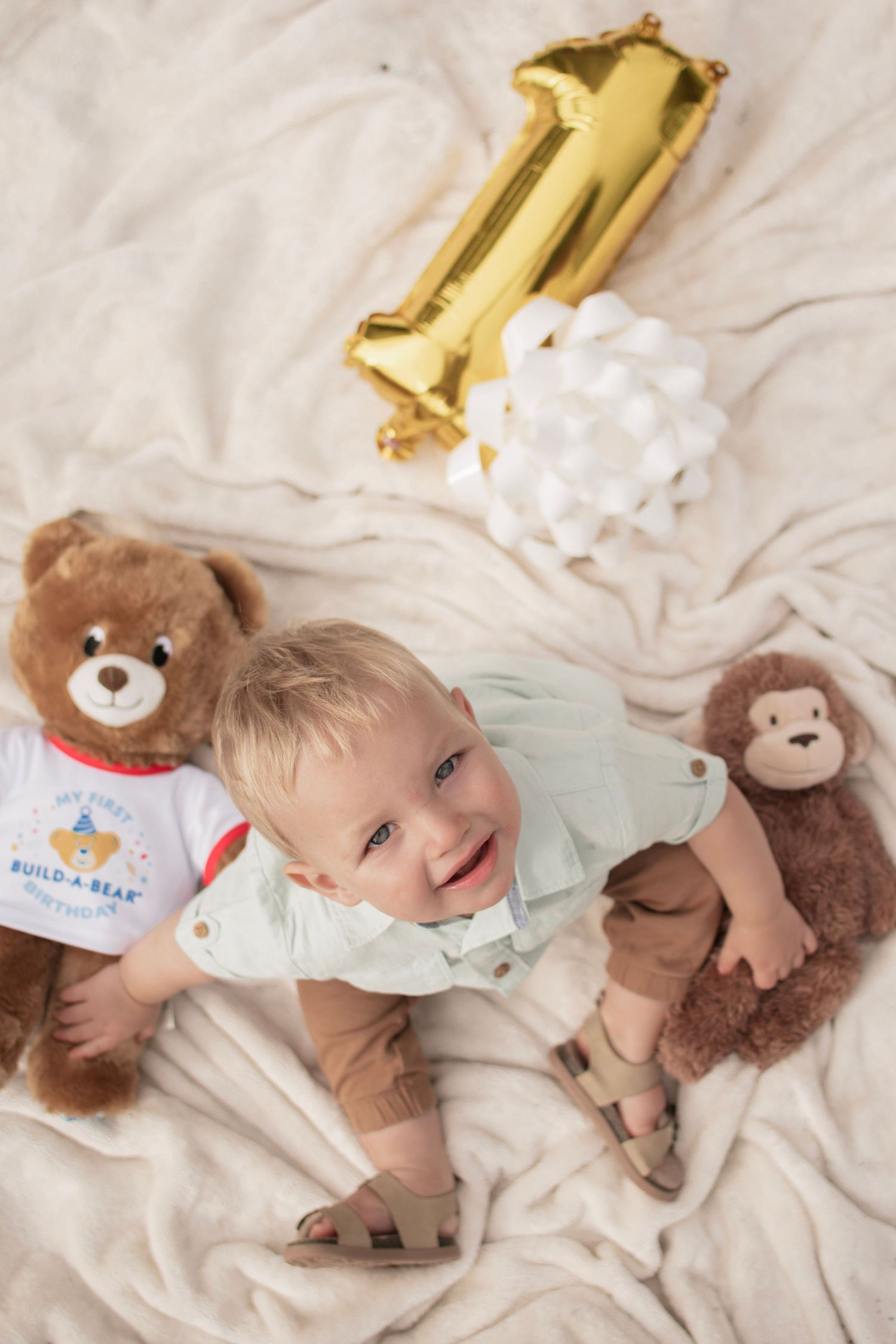 Baby smiling on blanket with teddy bears and gold number 1 balloon celebrating first birthday.
