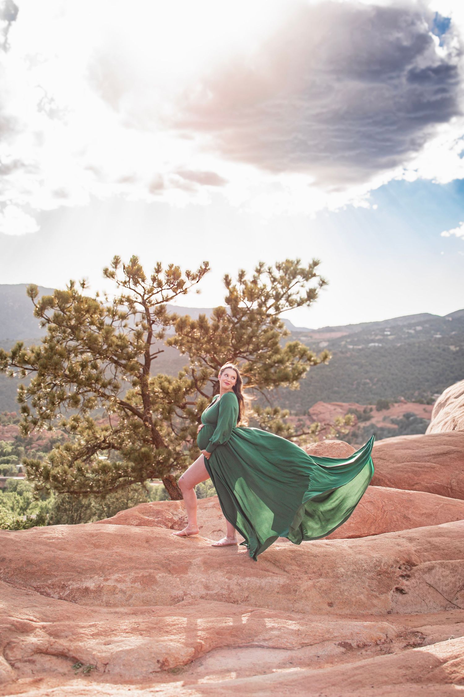 Pregnant woman in a flowing green gown poses on red sandstone rocks beside a pine tree under a dramatic sunset sky.
