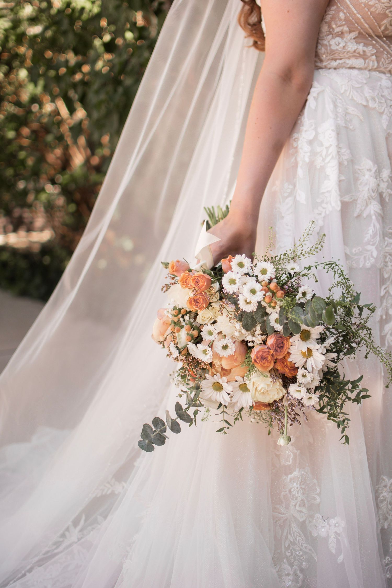 Bride in lace wedding gown holding a cascading bouquet of peach roses and white daisies with eucalyptus greenery.