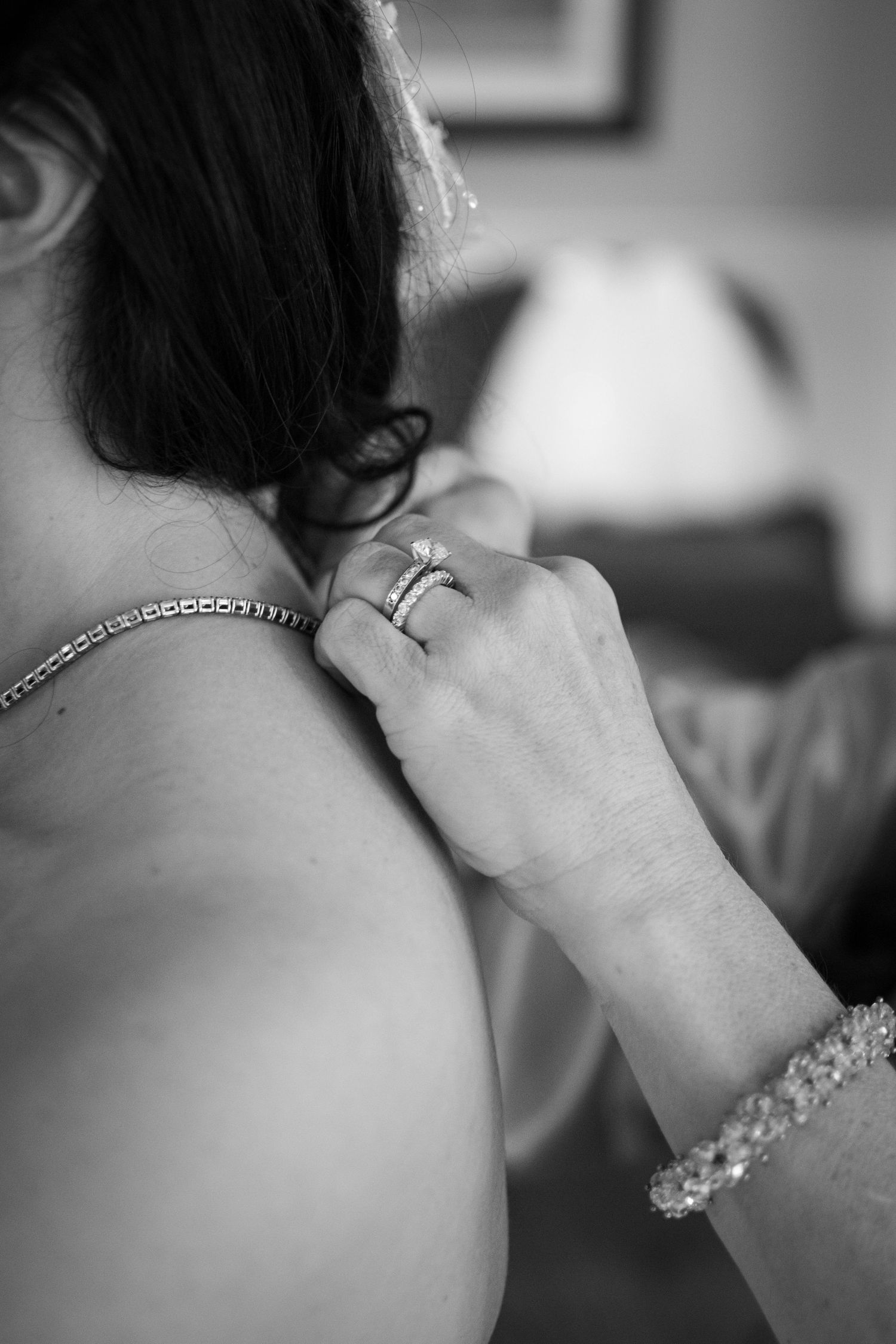 Black and white photo of a woman fastening a necklace on a bride, wearing diamond rings and a pearl bracelet.