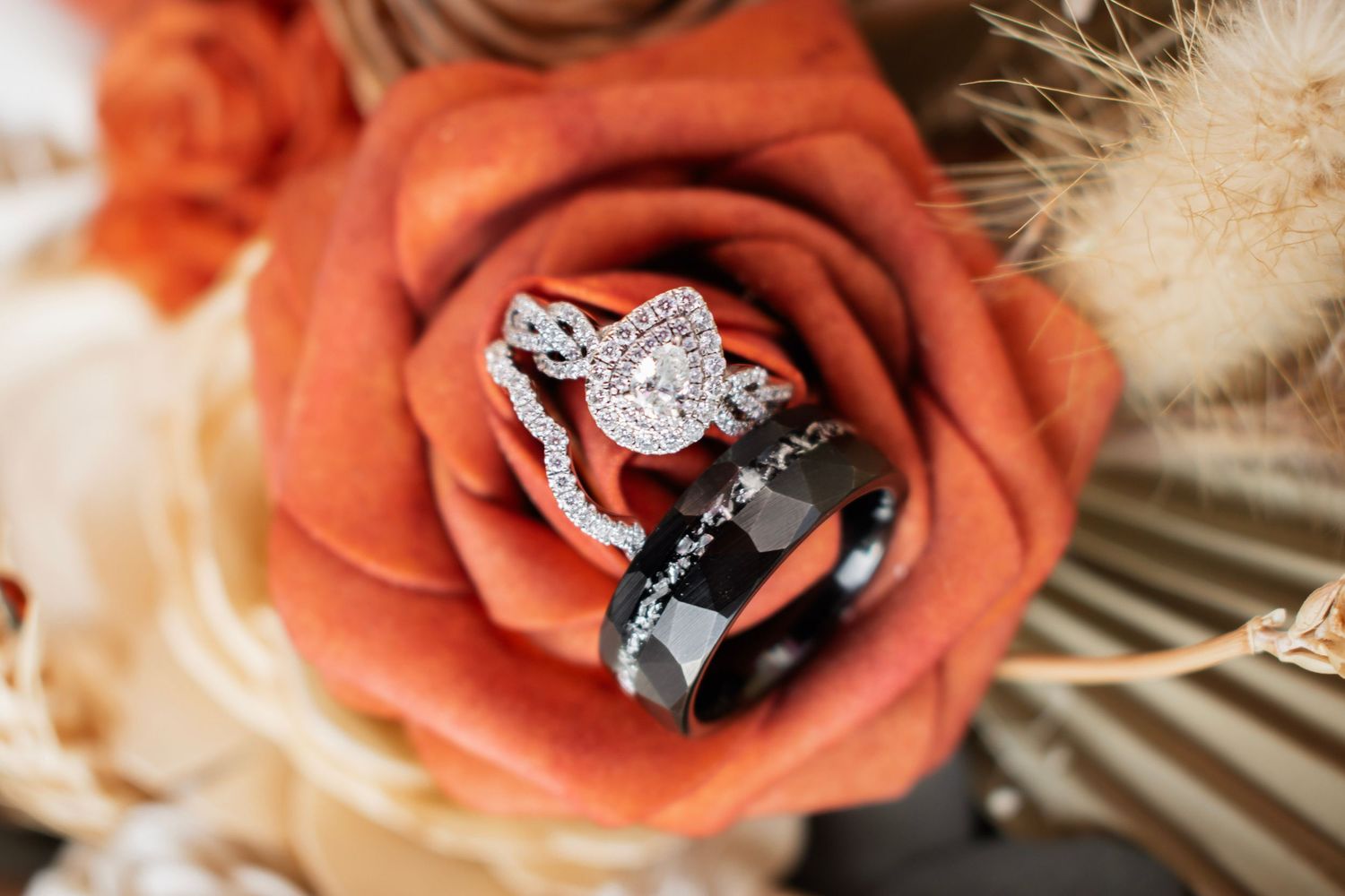 Wedding rings nestled in an orange rose, featuring a diamond halo engagement ring and black band in a floral bouquet.