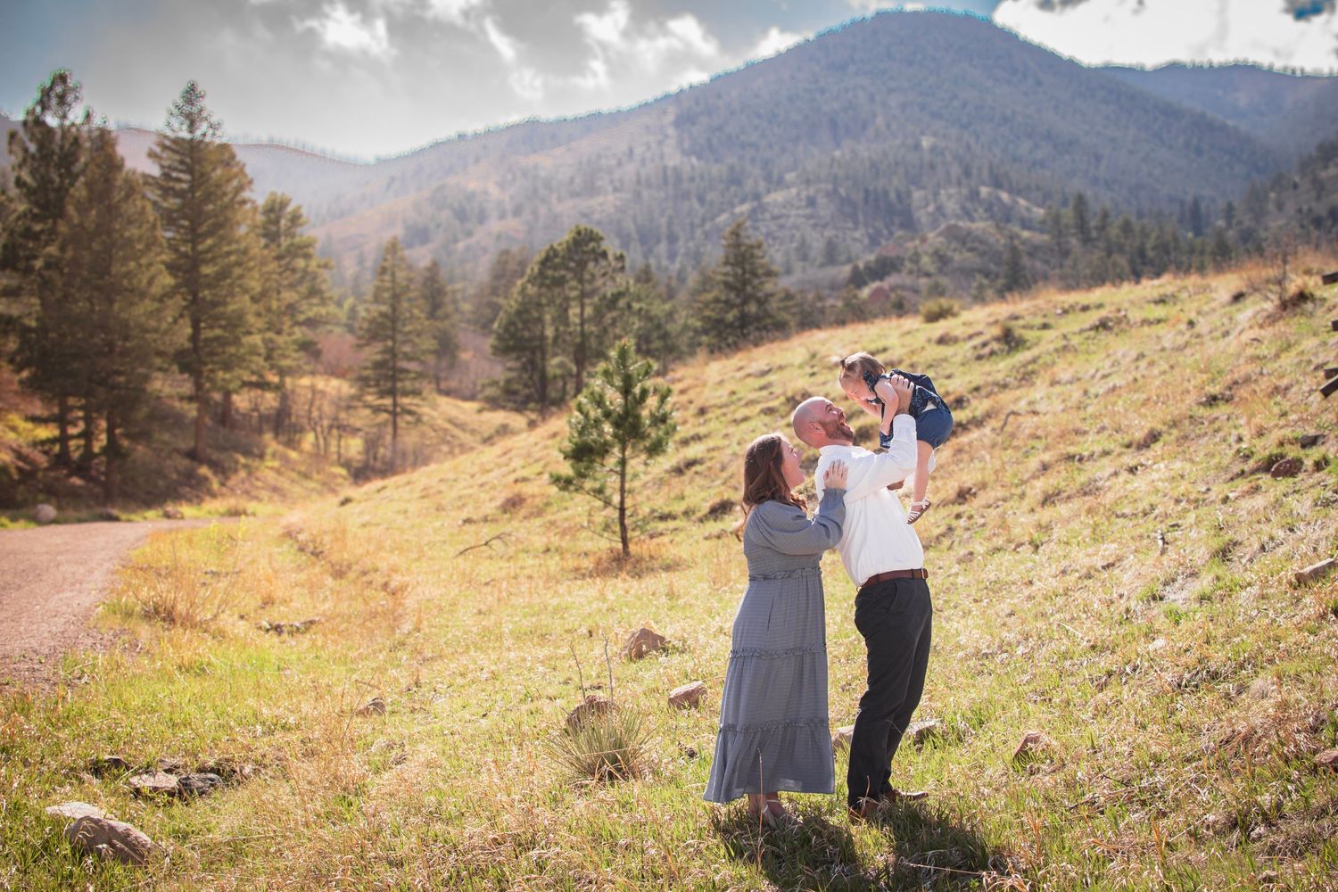 Family of three enjoying a sunny mountain meadow, parents lifting baby with pine trees and rocky hills in background at Blodgett open Space.