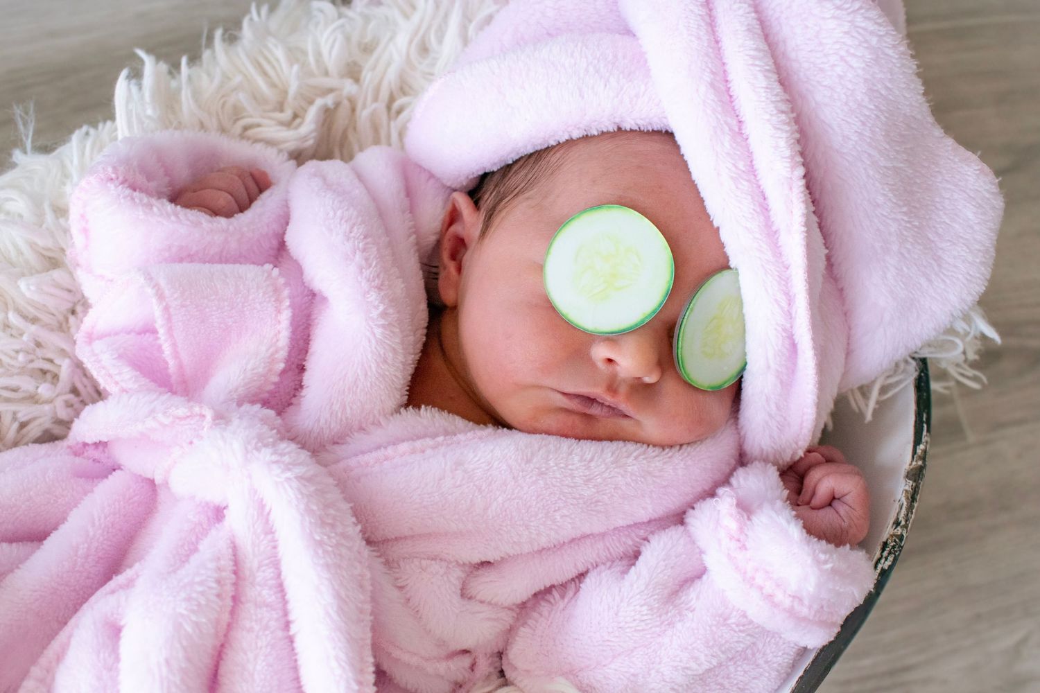 Newborn baby in pink bathrobe with cucumber slices on eyes, wrapped in fluffy towel, lying in basket spa treatment.