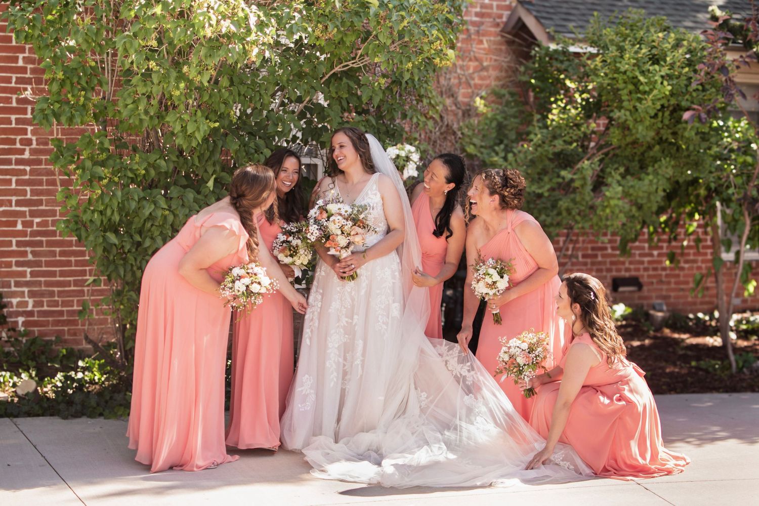 Bride in lace wedding gown surrounded by bridesmaids in coral dresses holding bouquets outdoors.