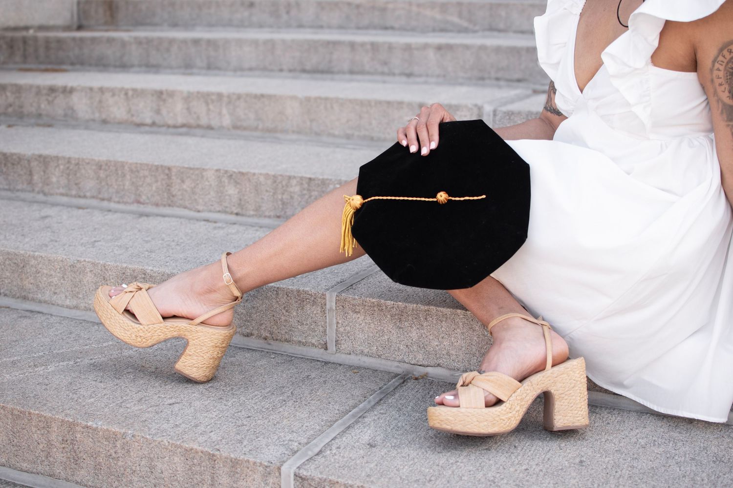 Woman in white dress wearing tan wooden platform heeled sandals, holding black graduation cap while sitting on stone steps.