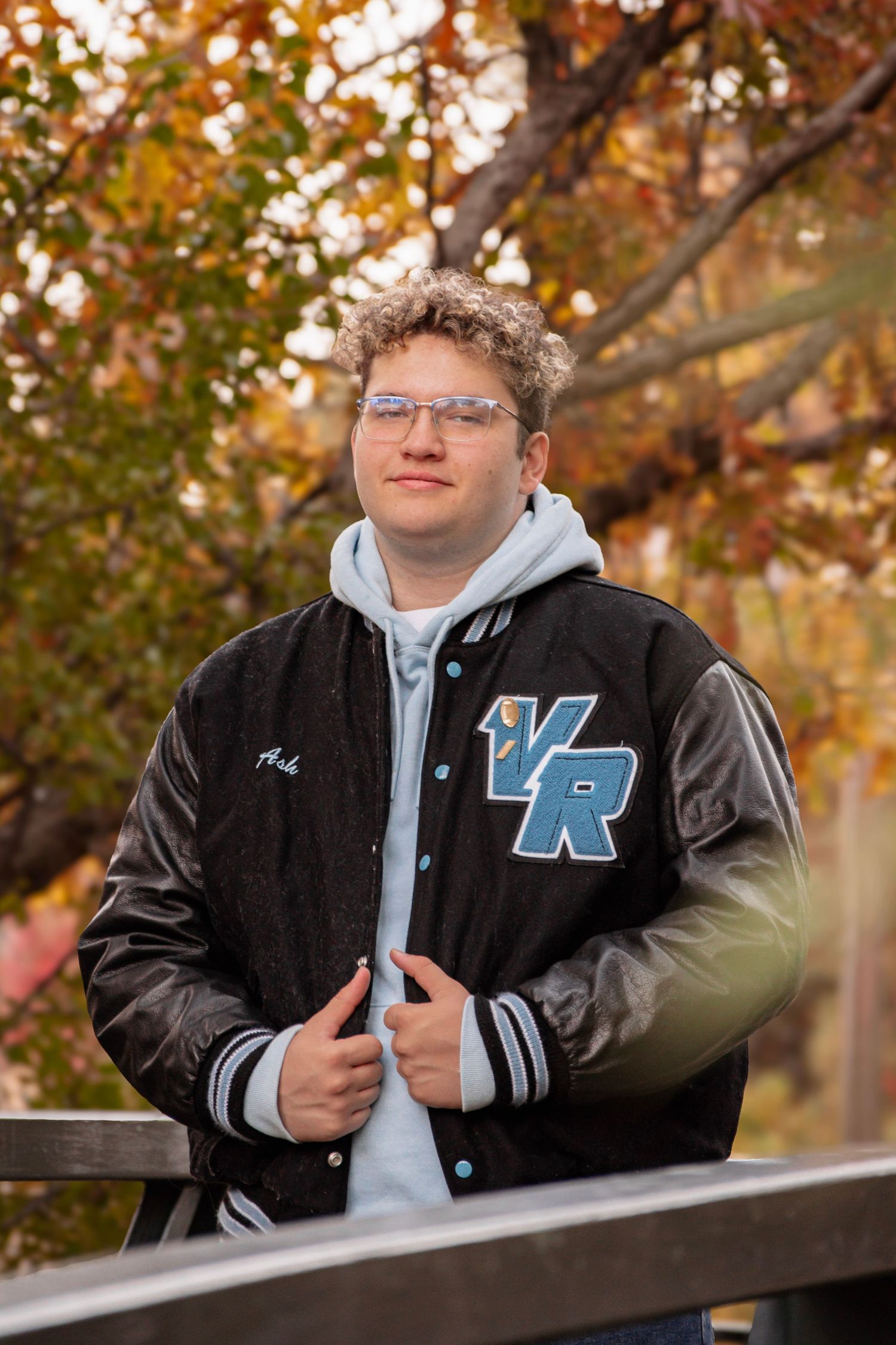 Young man in varsity jacket with curly hair and glasses poses outdoors among colorful autumn foliage.