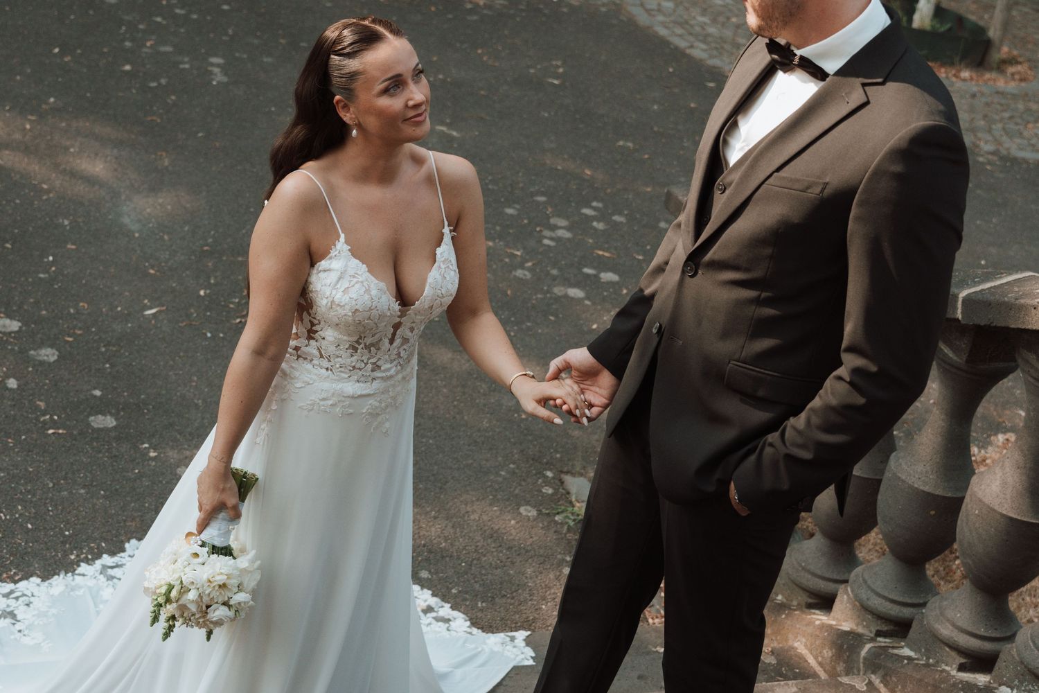 Bride in lace wedding gown holds bouquet while holding hands with groom in black suit.
