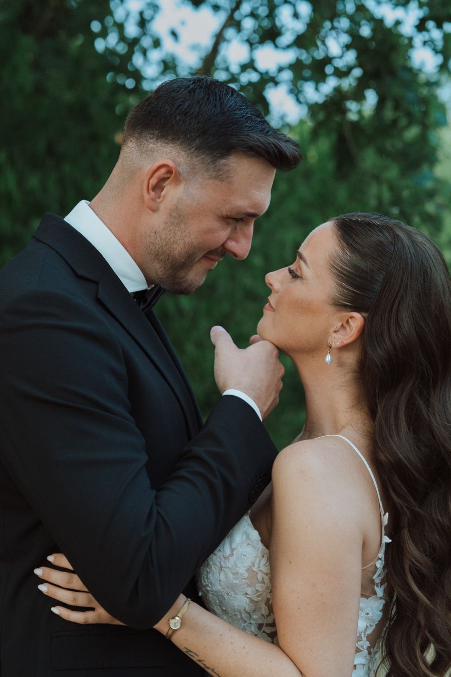 Groom in black tuxedo tenderly holding bride's chin in an intimate close-up wedding portrait outdoors.