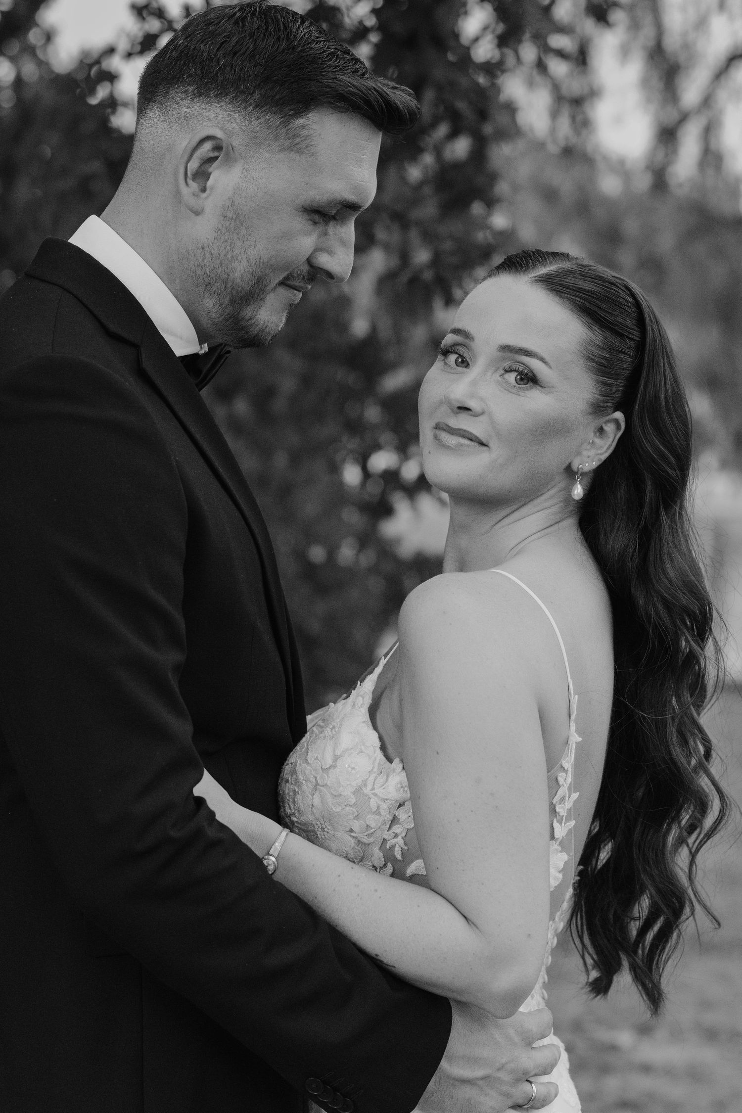 Groom tenderly holds bride as she gazes up at him in romantic black and white wedding portrait outdoors.