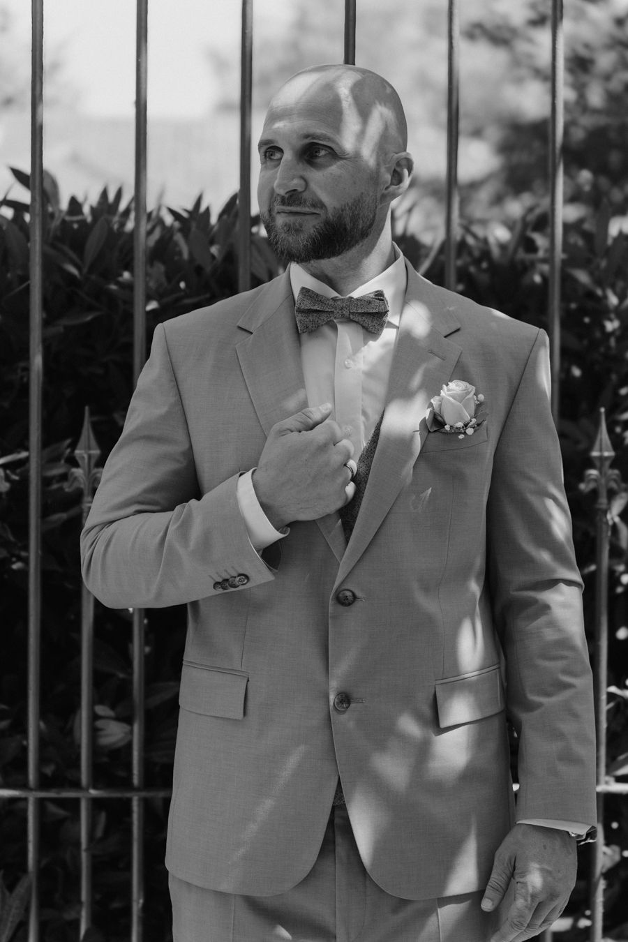 Bald groom in gray suit with bow tie and boutonniere poses by iron fence.