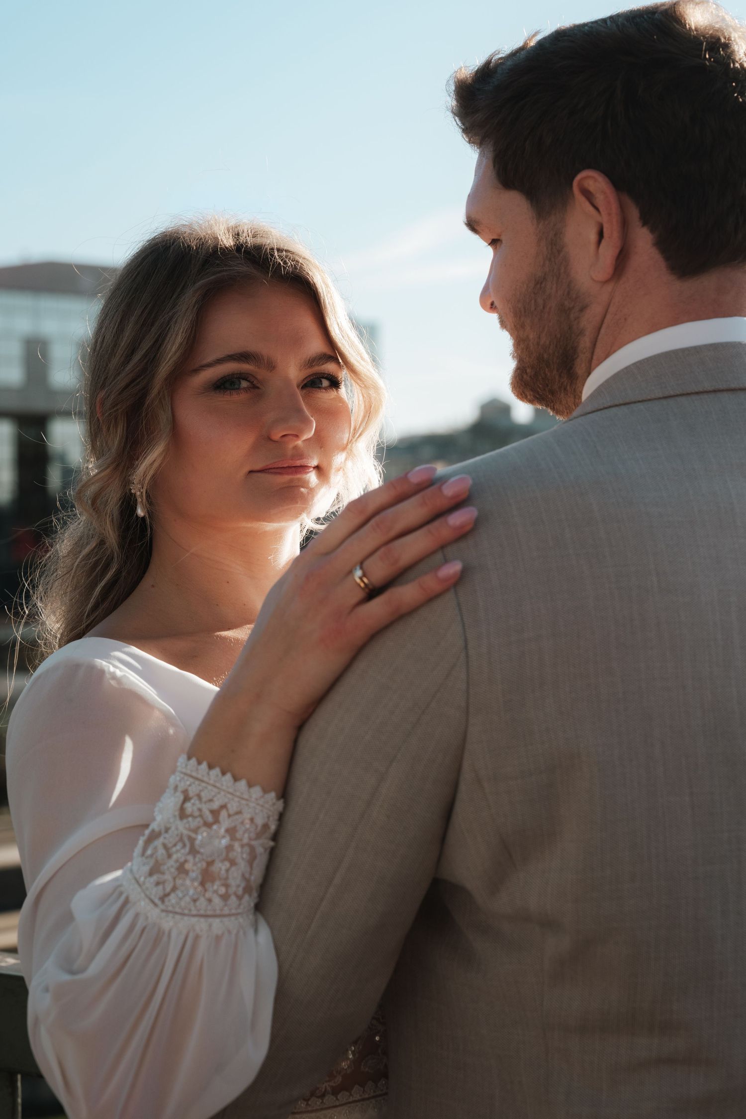Bride in white lace dress places hand on grooms gray suit shoulder during outdoor wedding photo.