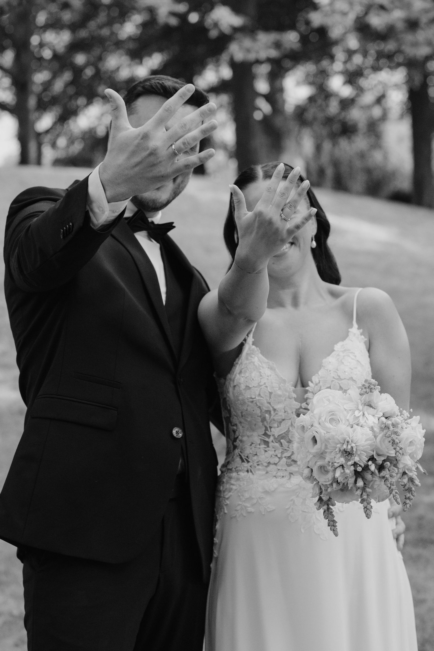 Bride and groom playfully hiding faces with hands showing wedding rings in black and white outdoor photo.