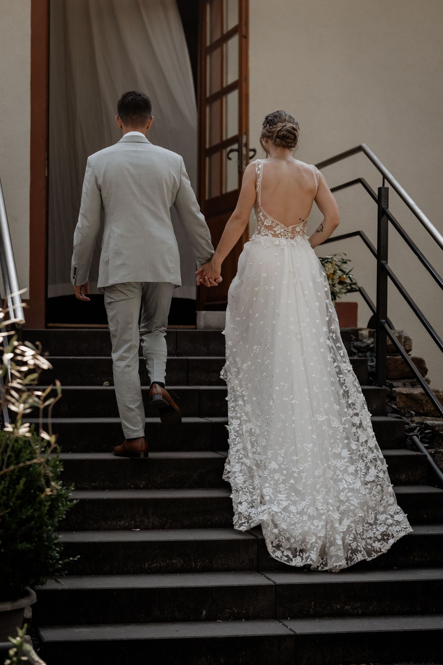 Bride and groom walking up stone steps hand in hand, bride wearing a backless lace gown.