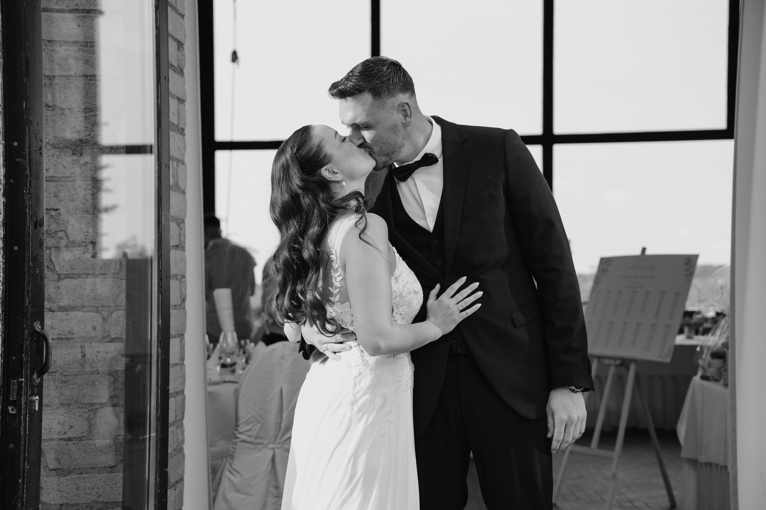 Bride and groom sharing a kiss in black and white photo near industrial glass doors at wedding venue.