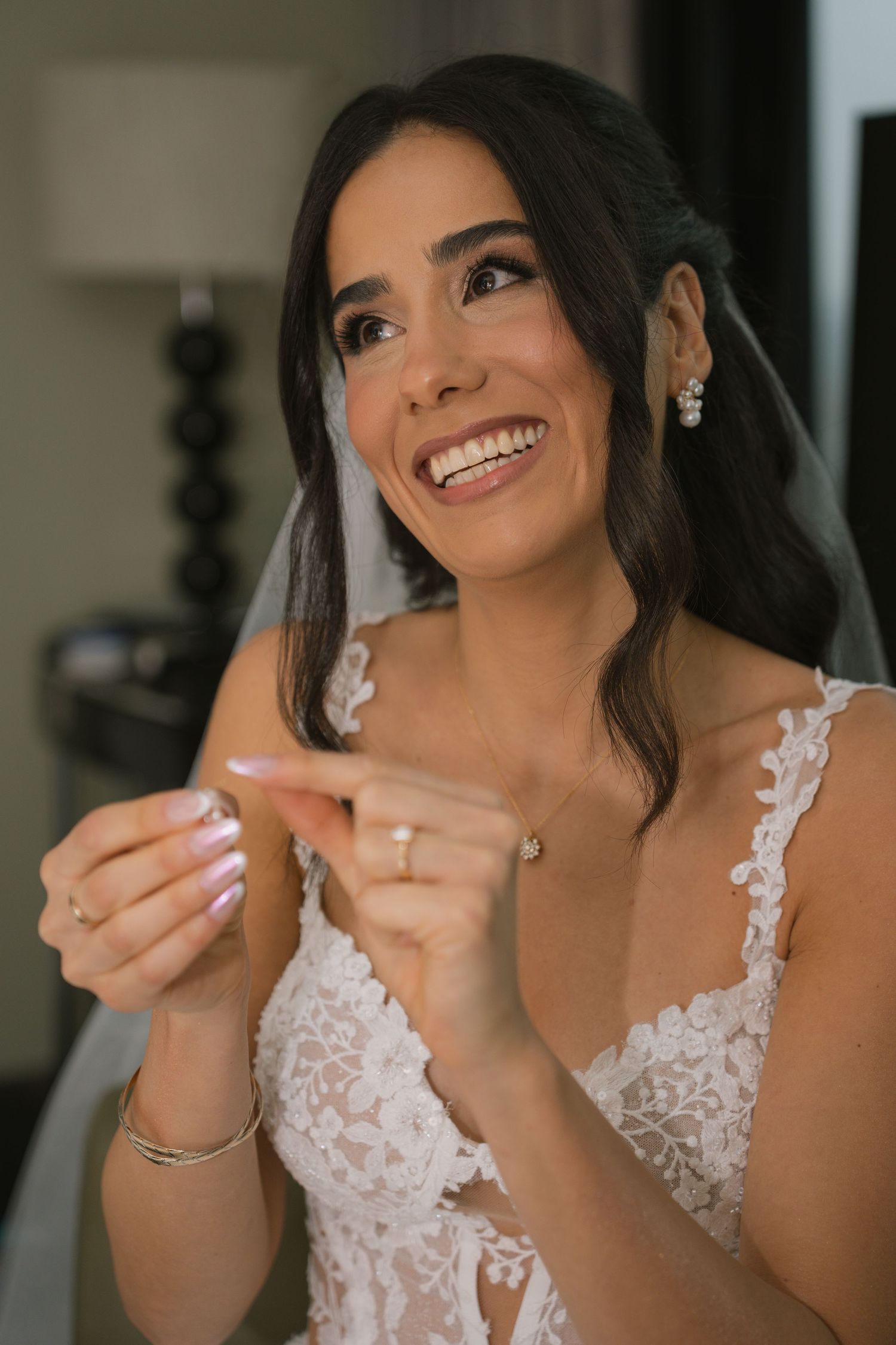 Radiant bride in lace wedding gown smiling joyfully while adjusting her ring, wearing pearl earrings and veil.