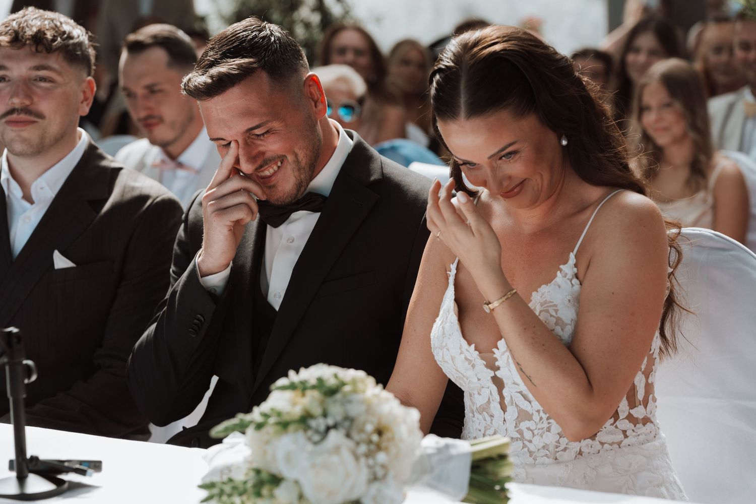 Emotional bride and groom wiping tears during heartfelt wedding ceremony at decorated table.