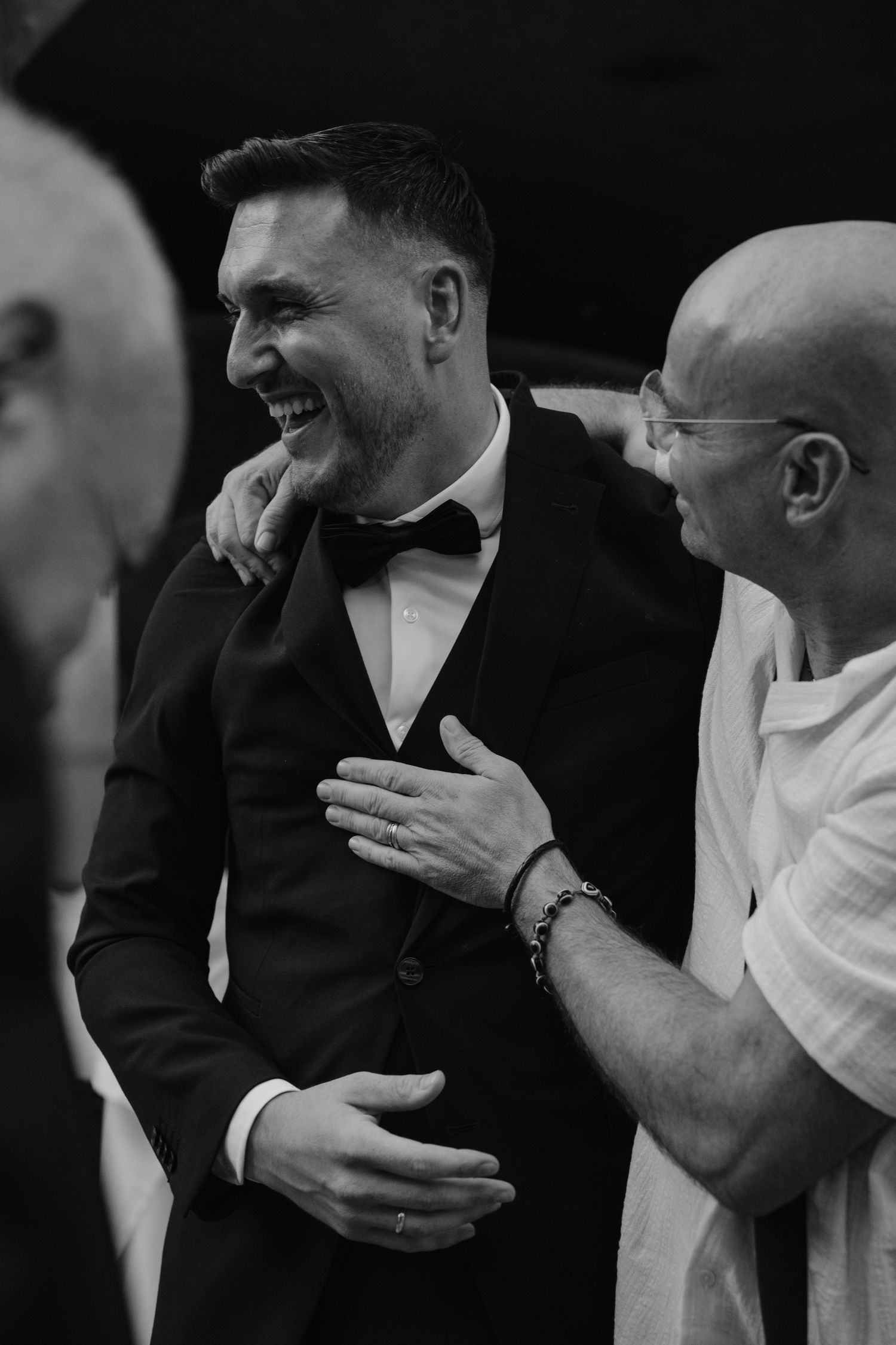 Groom in black tuxedo laughing joyfully with guests at wedding reception in black and white photo.